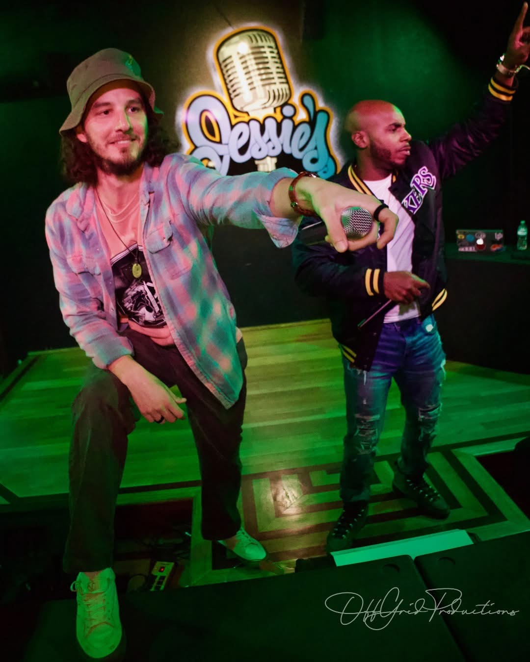 Two men perform on stage with a microphone and a DJ setup, colorful lighting, and a sign reading 'Jessie' in the background.