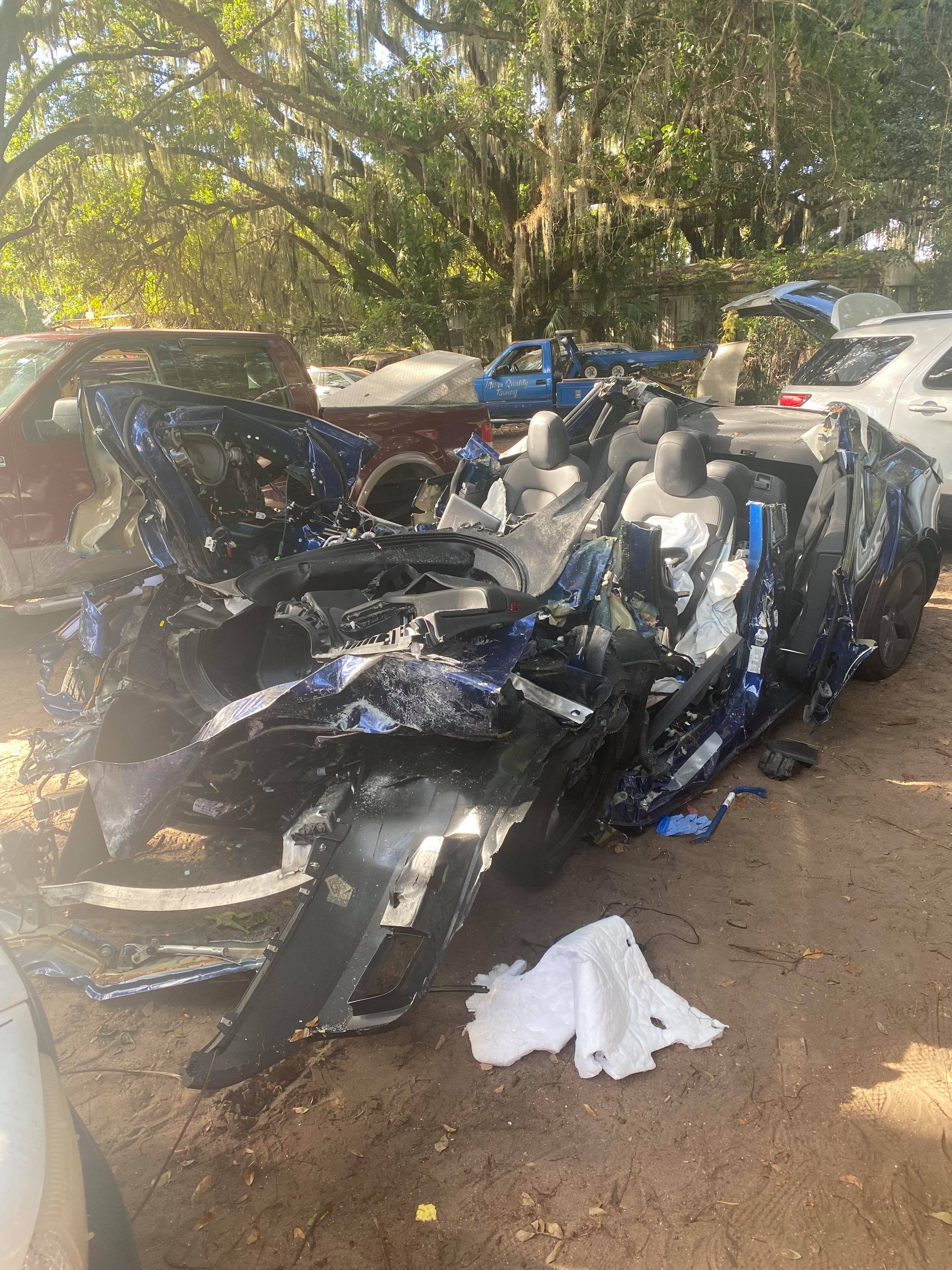 Damaged motorcycle in a lot with other vehicles and trees in the background.