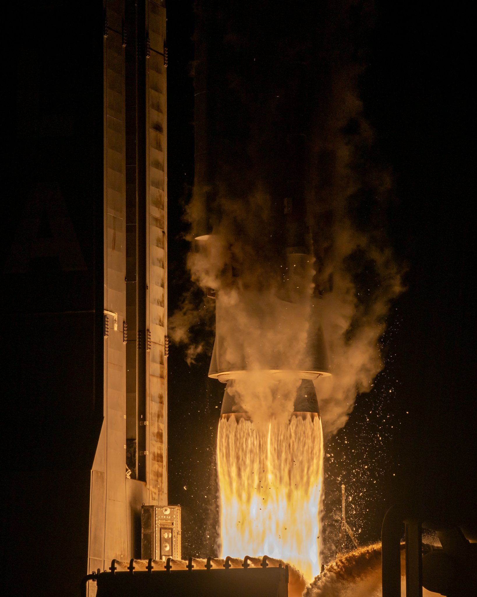 Industrial process with molten material pouring from a large container, emitting bright light and smoke at night.