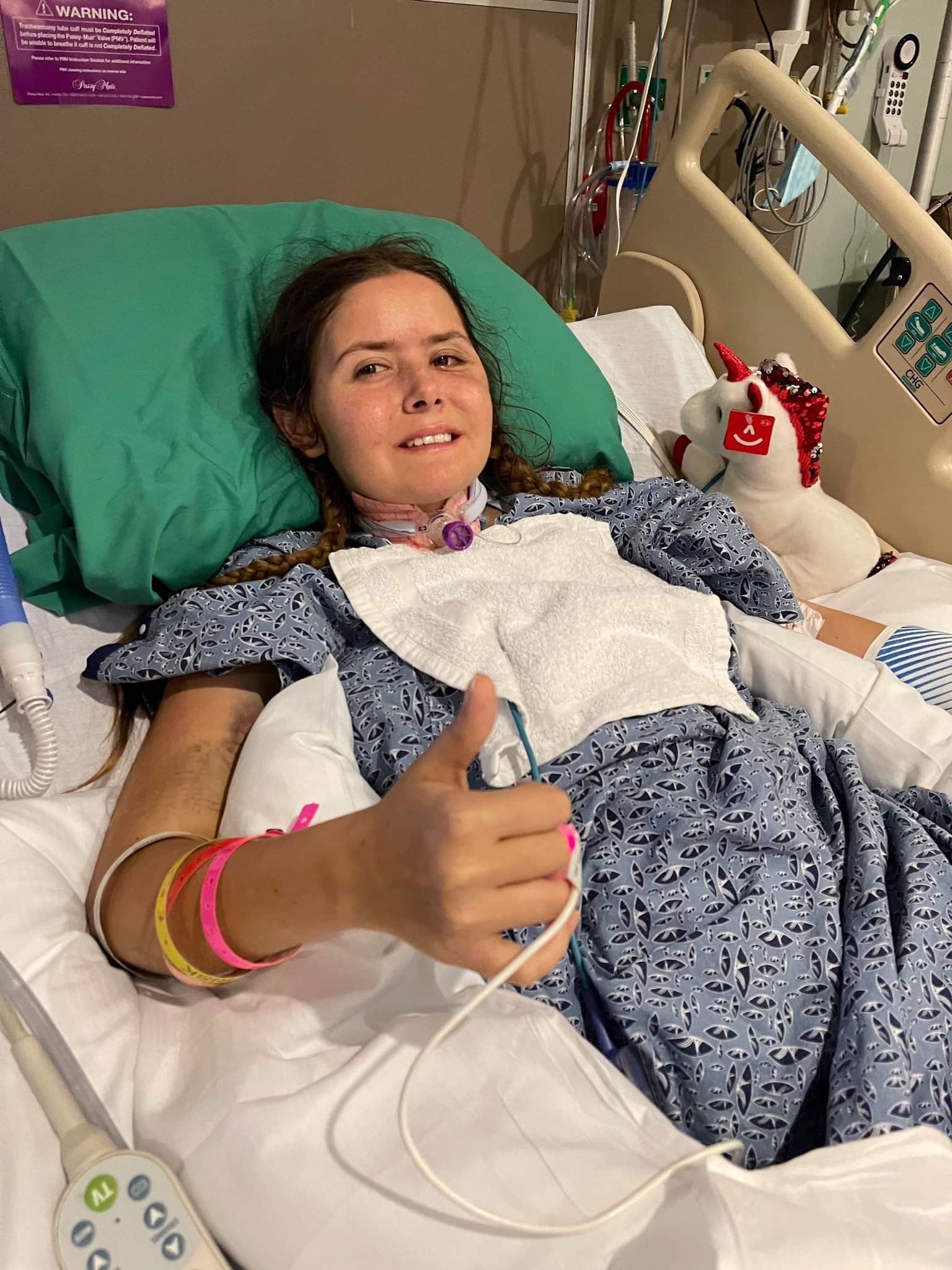 Young girl lying in hospital bed giving a thumbs-up, wearing hospital gown and wristbands, with medical equipment nearby.