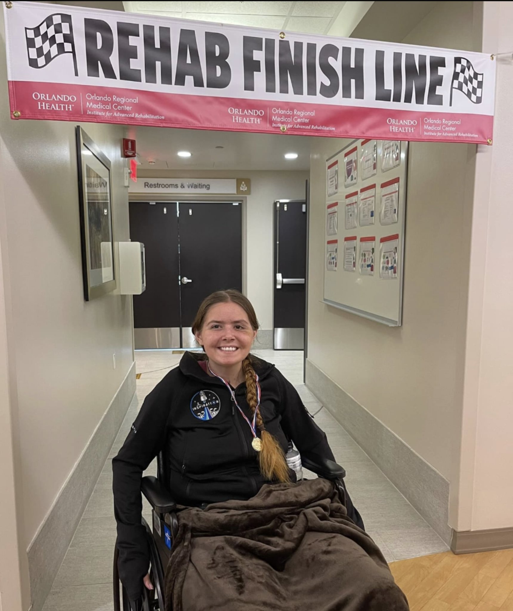 Smiling woman in a wheelchair at rehab finish line with a pink banner overhead, hallway background, and bulletin board on the wall.