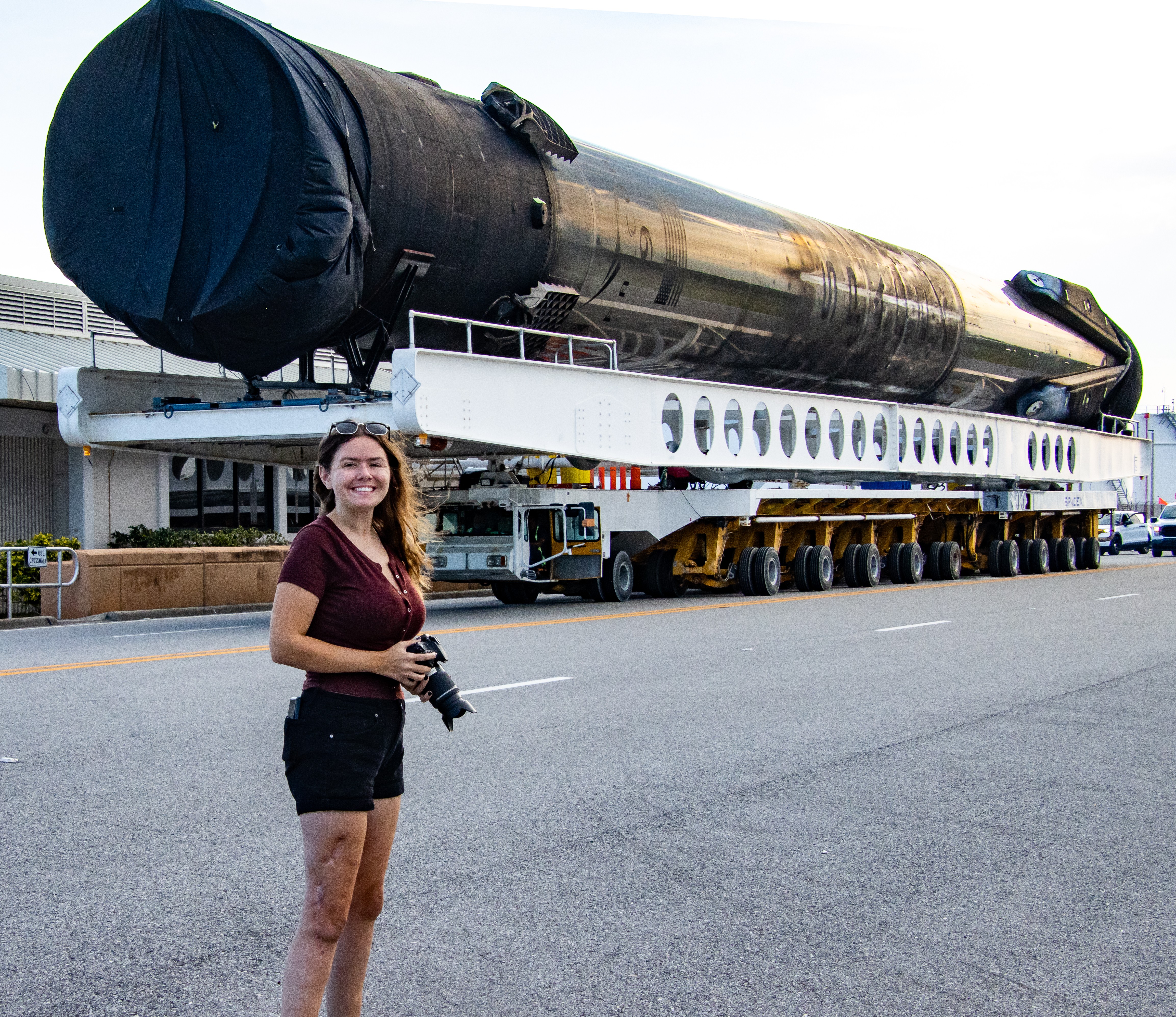 Young woman holding a camera standing in front of a large rocket on a transport vehicle, outdoors.