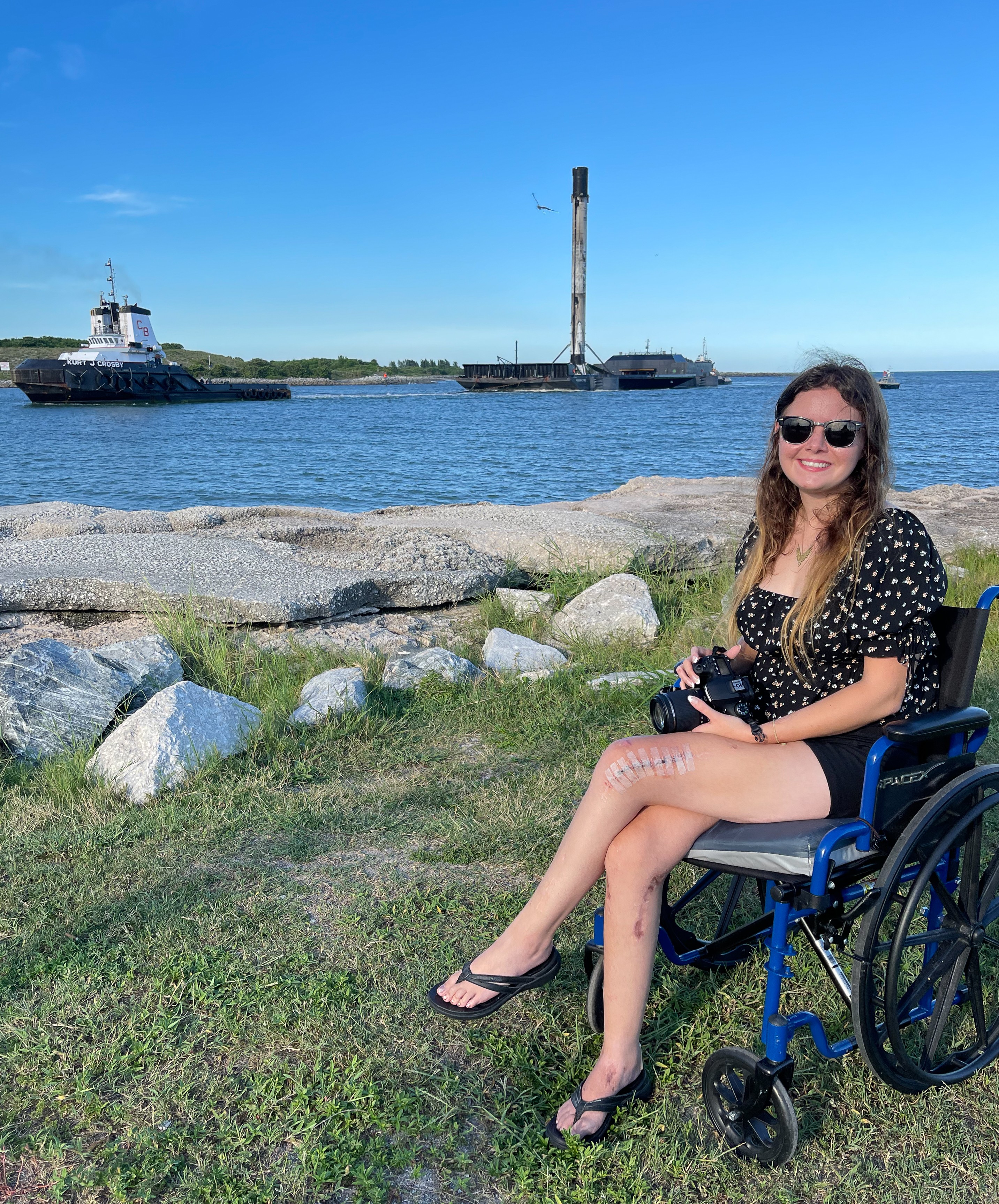 Young woman in sunglasses sitting in a wheelchair on grass near water with boats and a lighthouse in background.