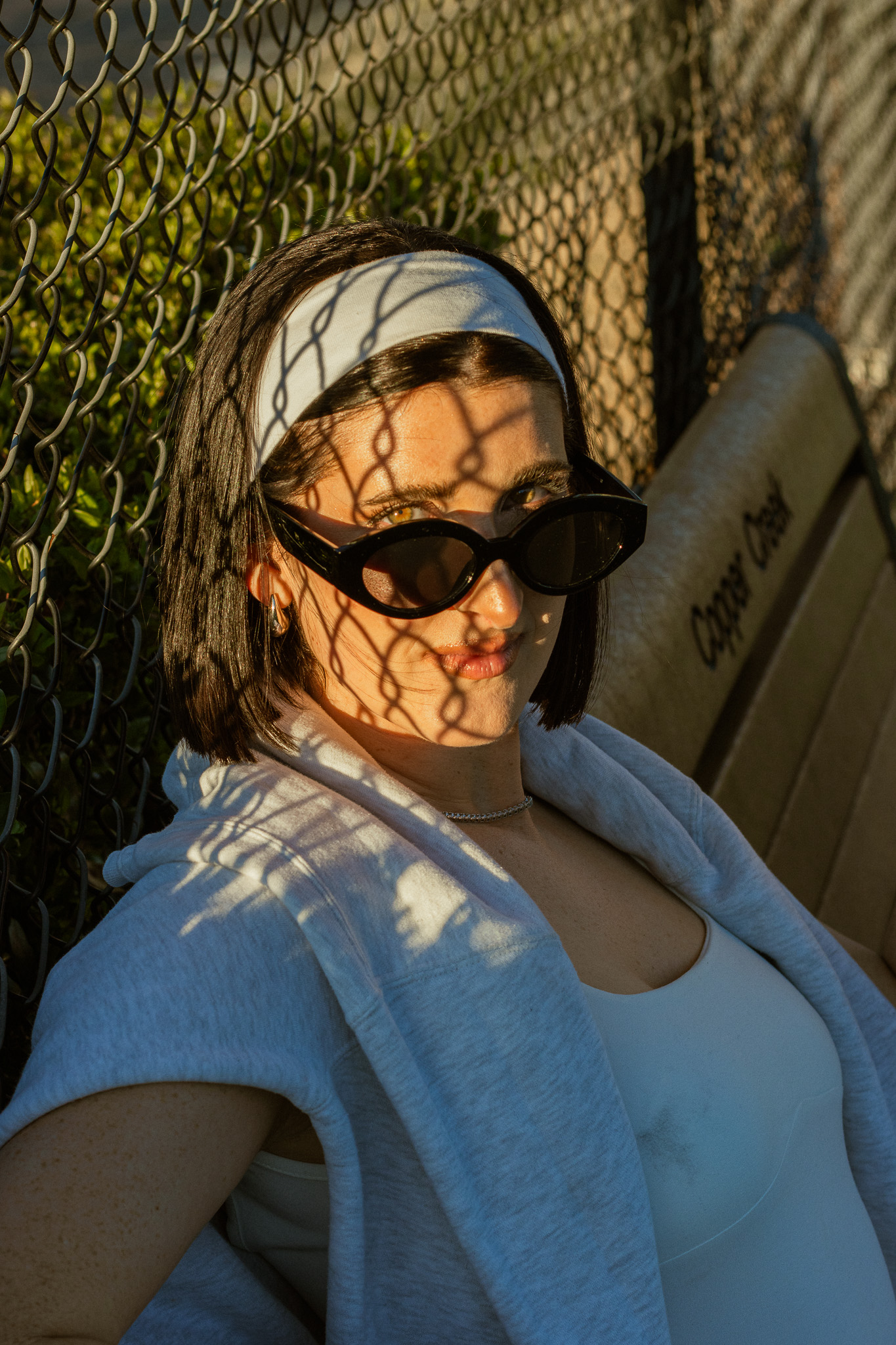 Woman with sunglasses and headband sitting on a bench, shadows from a chain-link fence on her face and background.