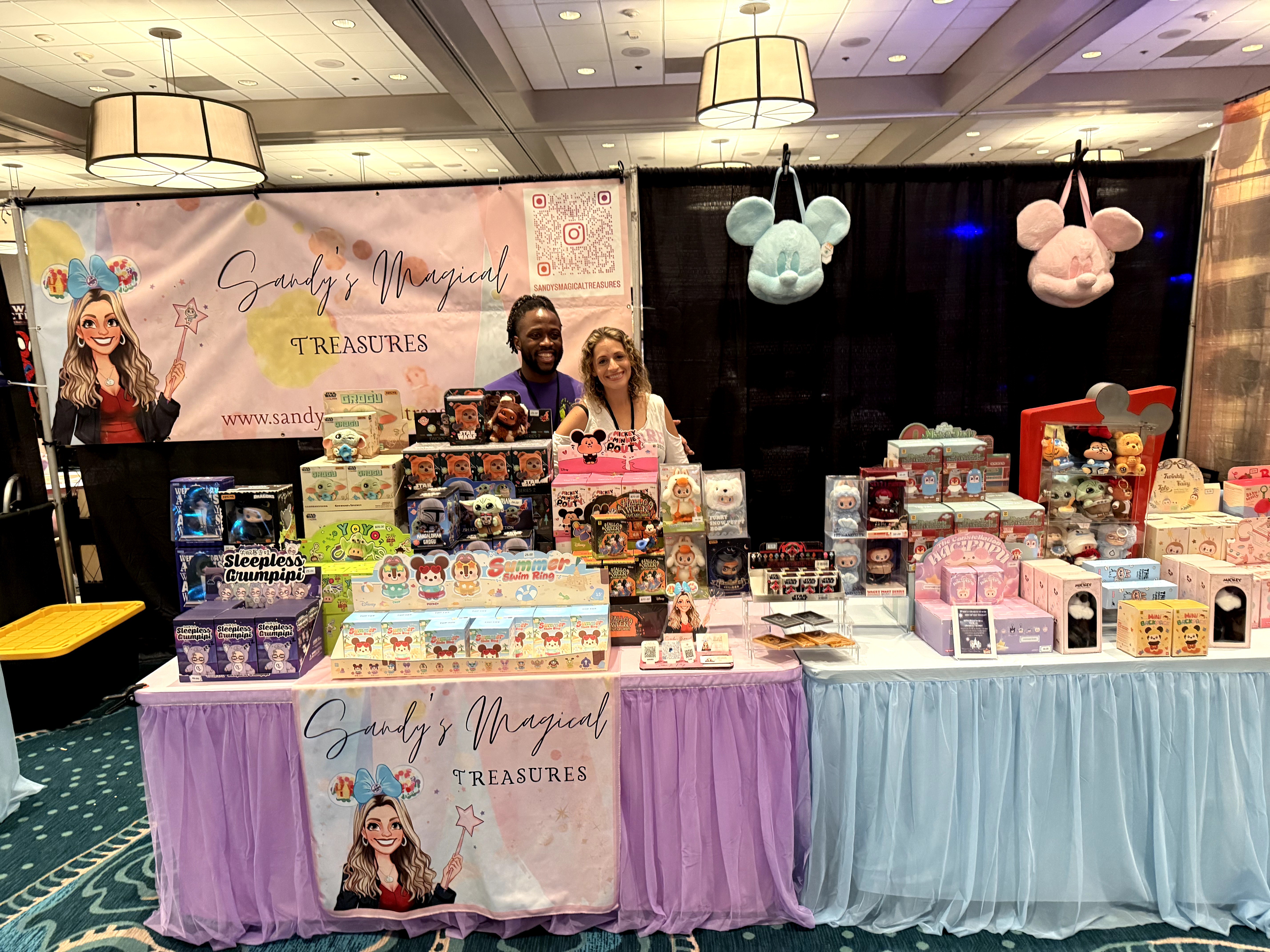 Two people stand behind a table filled with colorful merchandise, with a banner and hanging plush toys in the background.