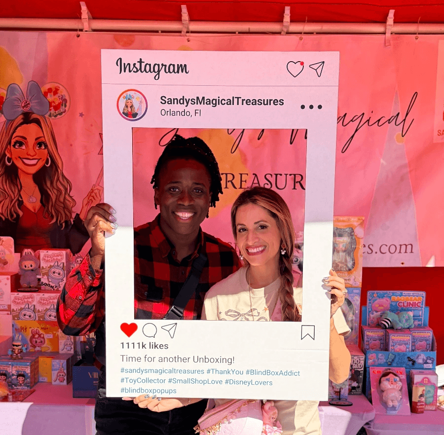 Two women smiling behind a large Instagram frame prop at an event, with pink background and merchandise displays.