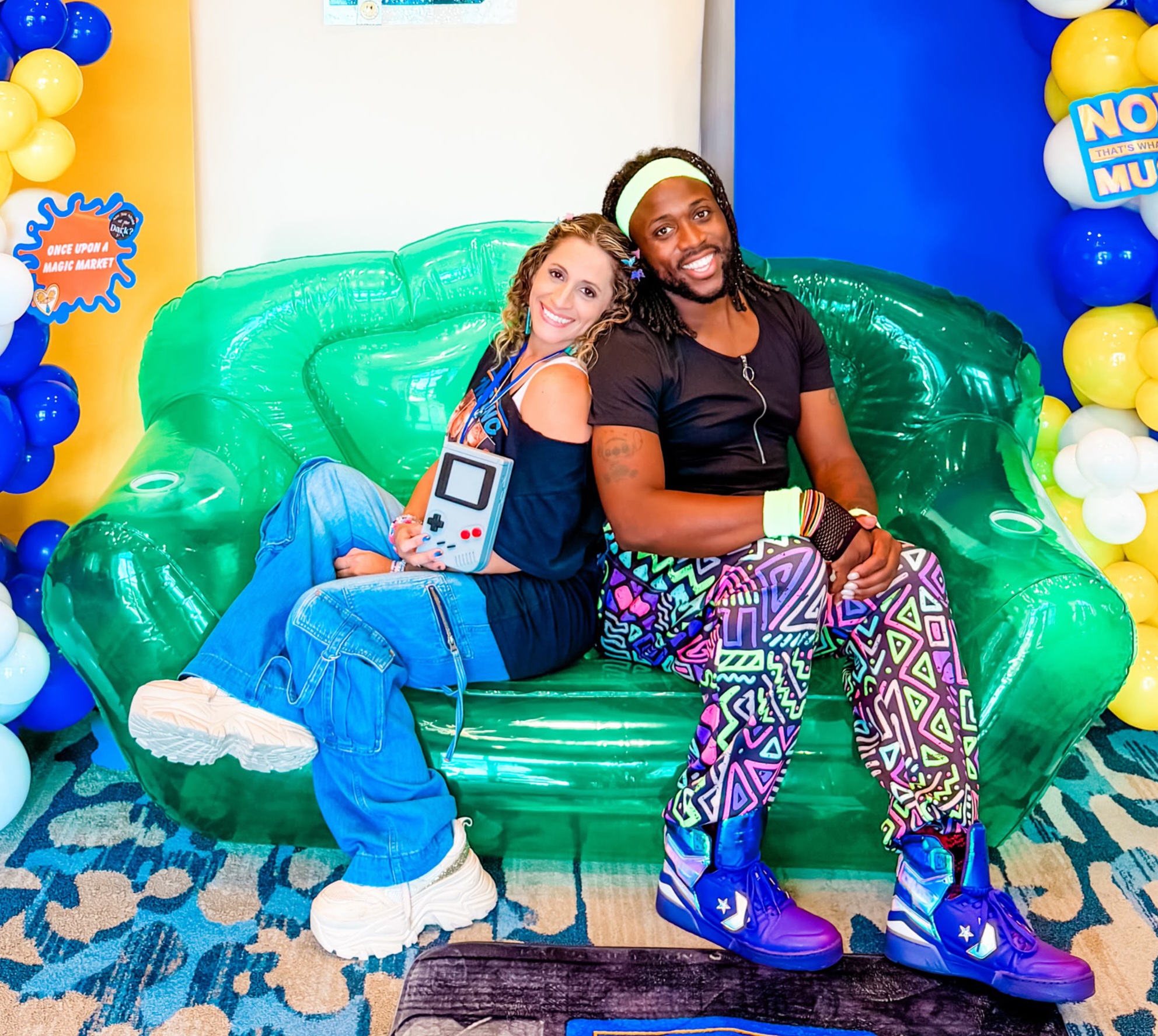Two people sitting on a green inflatable couch, smiling, with colorful balloons and decorations in the background.