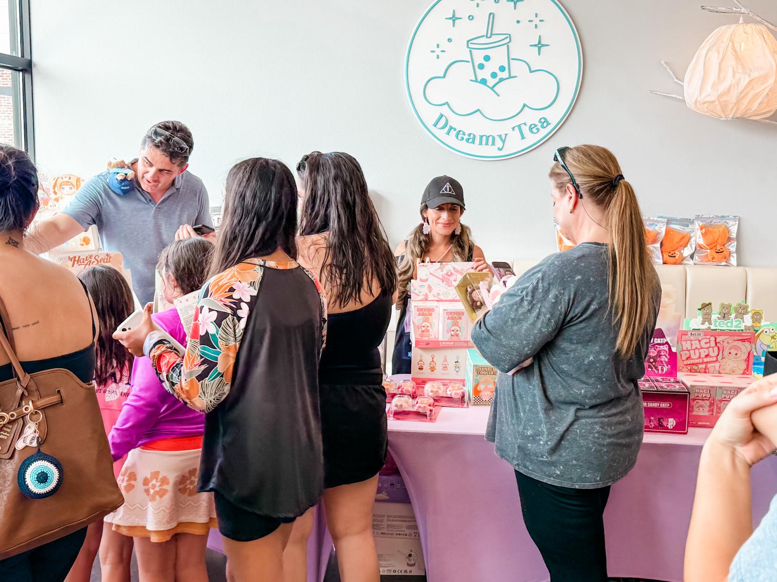 Group of people gathered around a table at a tea party event, with a sign reading 'Dreamy Tea' on the wall.