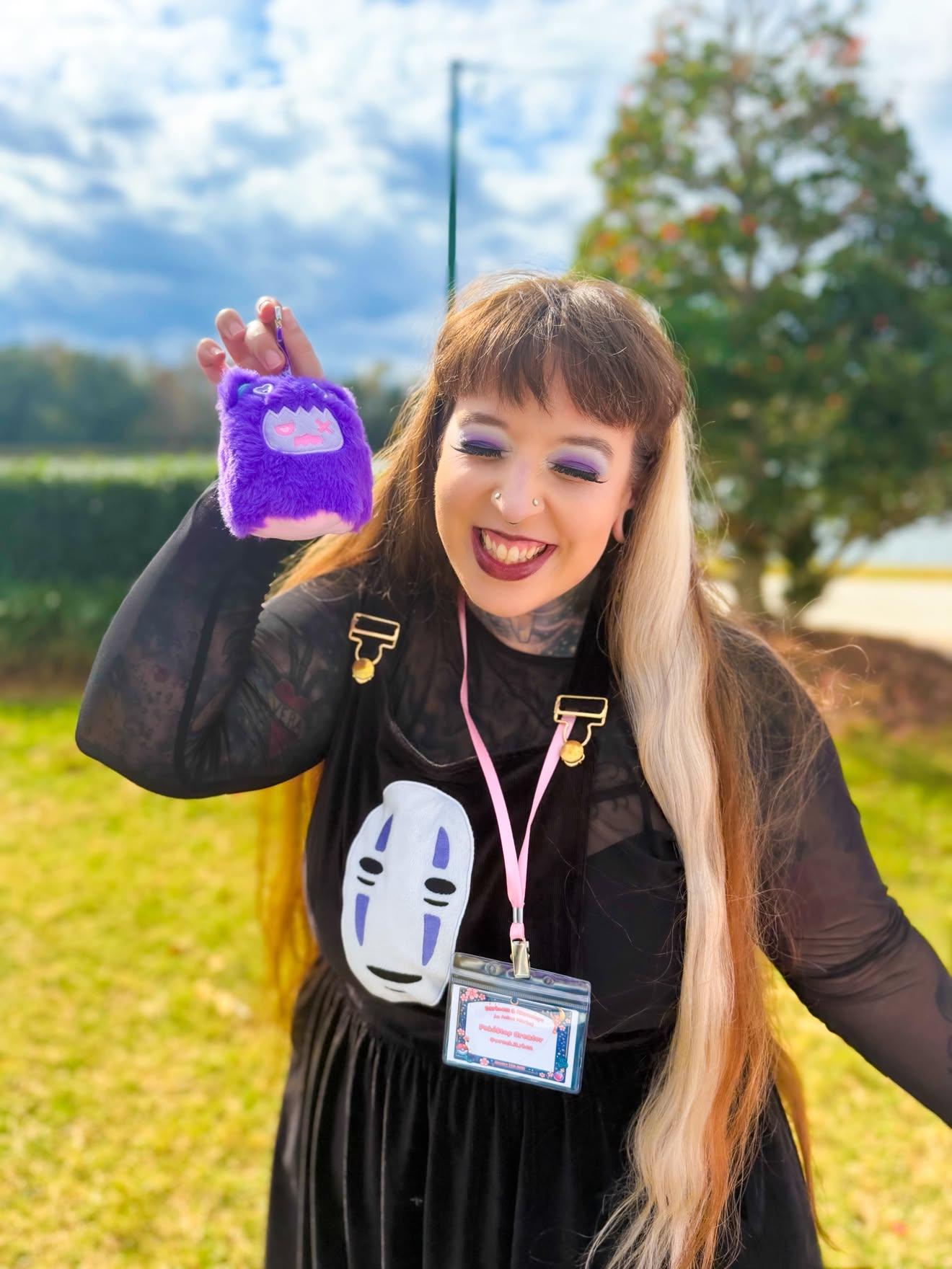 Young woman outdoors smiling, holding a purple plush bag, wearing a black dress, with a name badge and pink earphones, trees and sky in background.