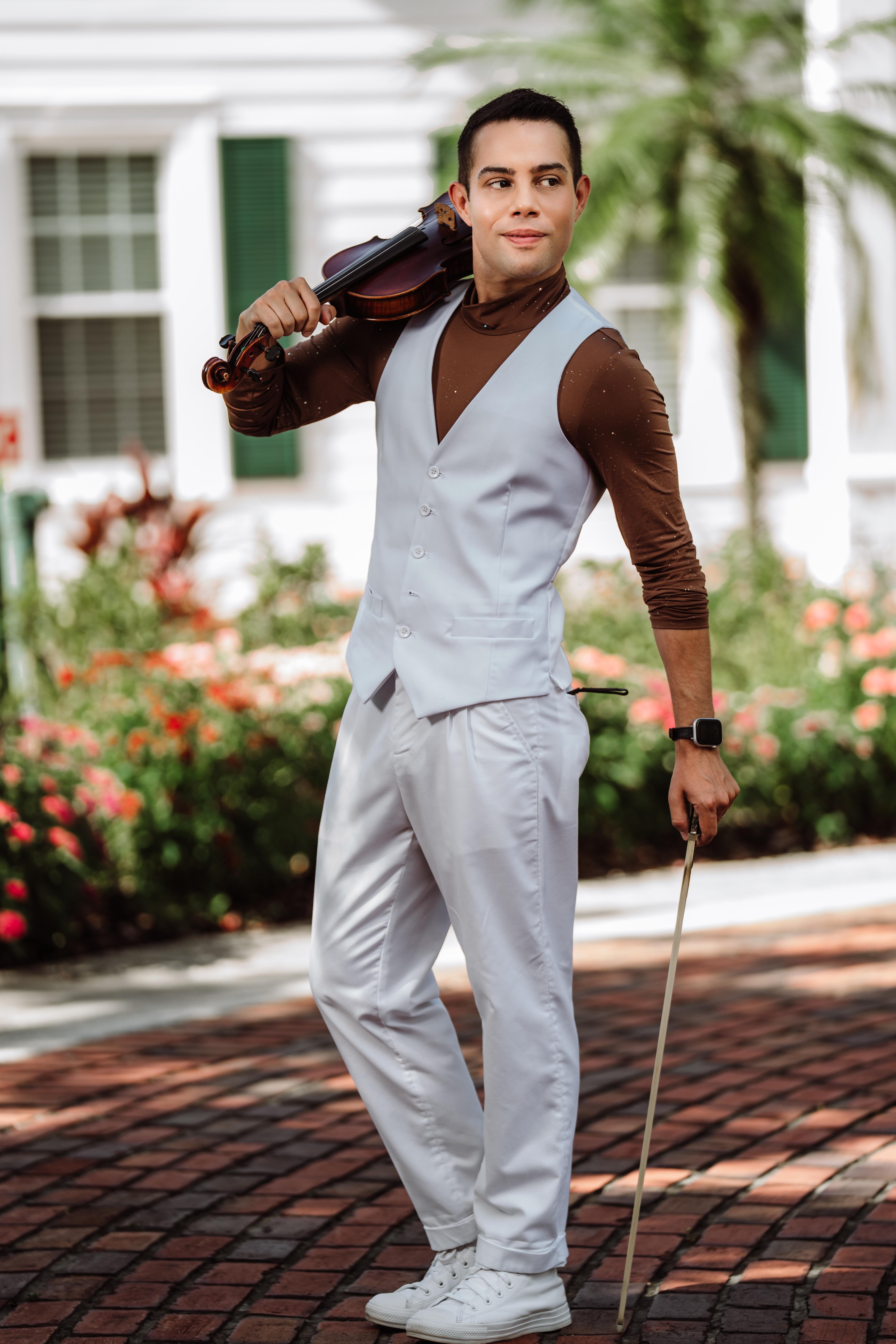 Young man in light gray suit holding a violin and bow outdoors, with greenery and a white house in background.