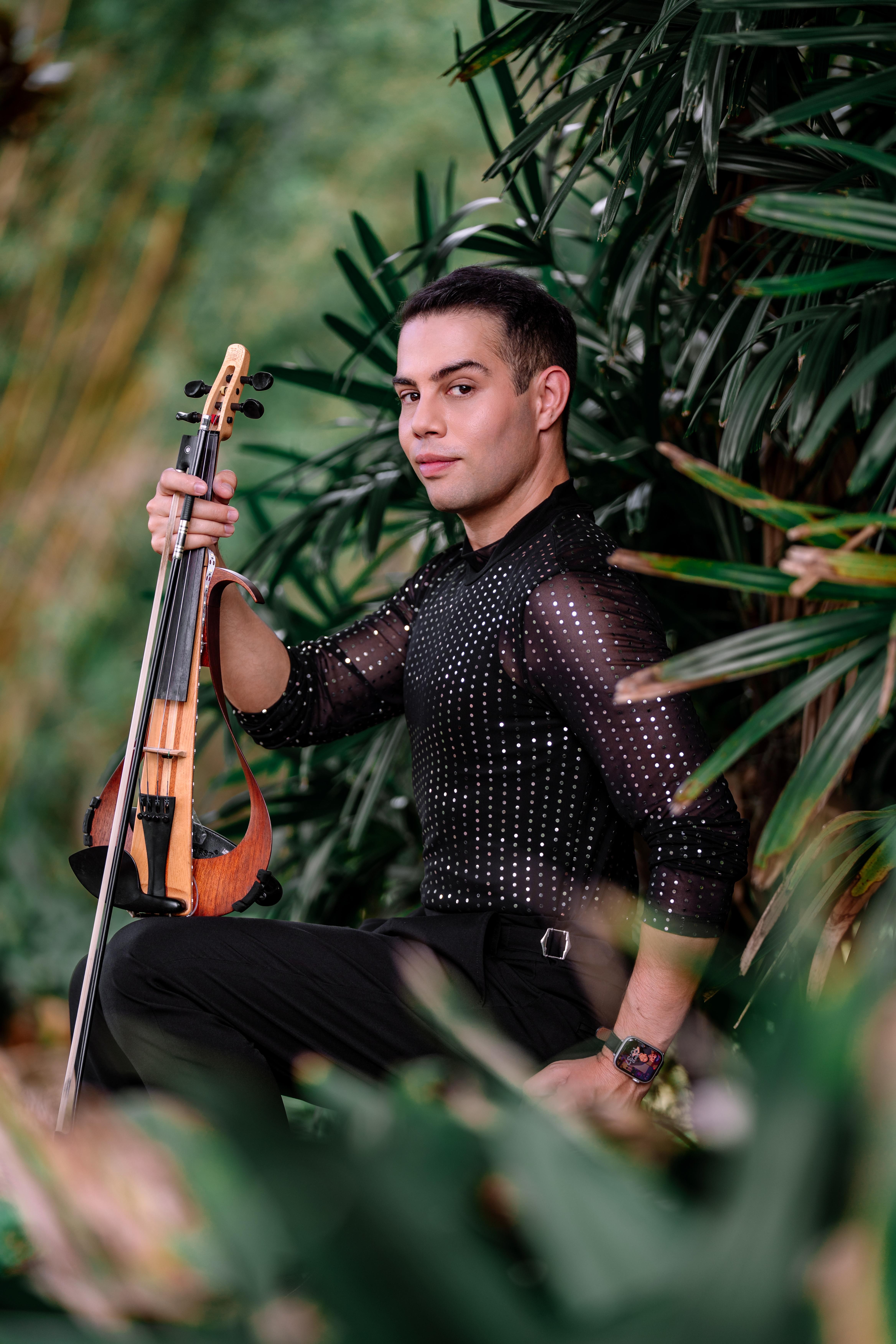 Man sitting outdoors among lush green plants, holding a guitar, wearing a black, semi-transparent, dotted shirt, looking to the side.