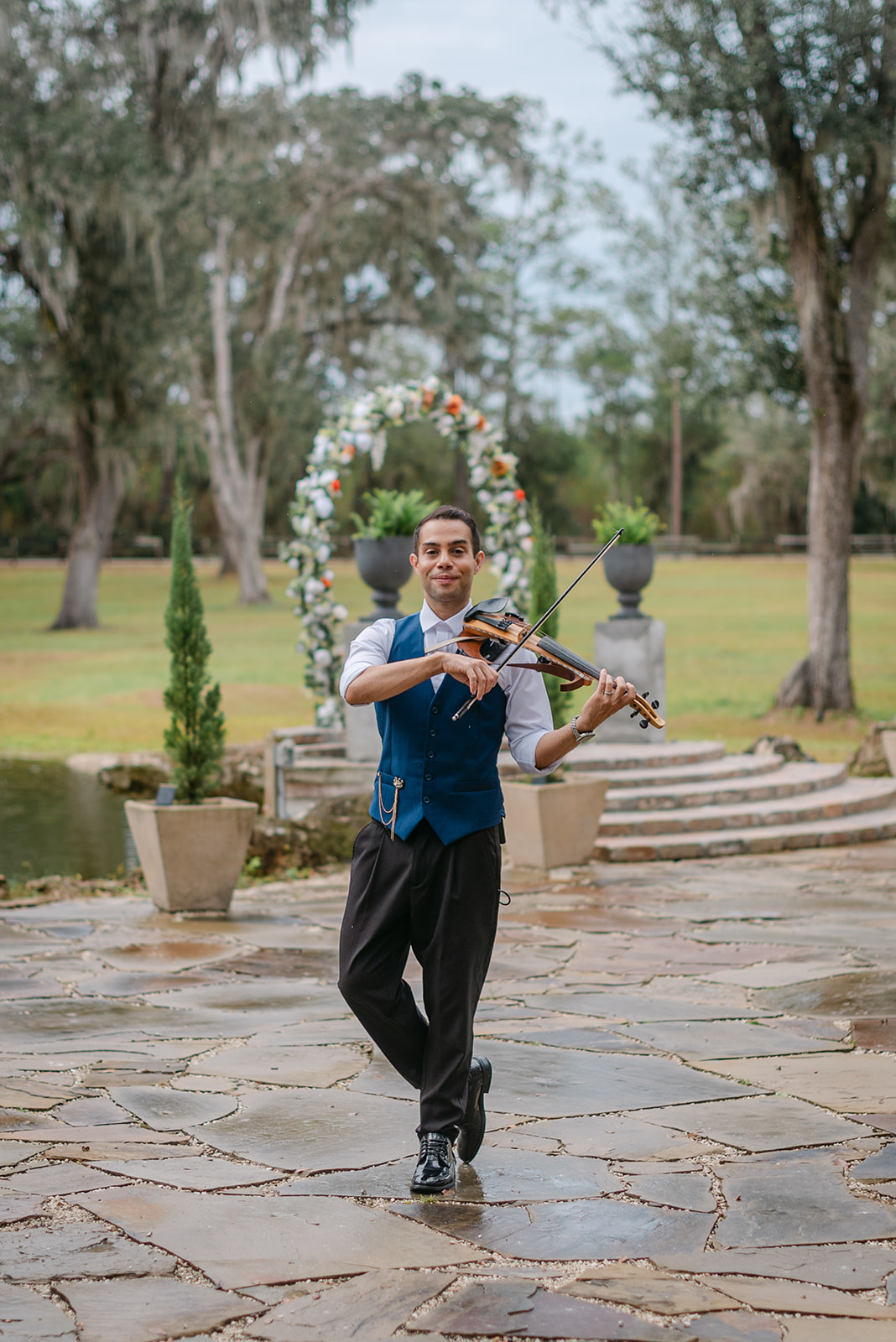 Young man playing violin outdoors in a park with trees and stone pathway, smiling, wearing a blue vest and black pants.