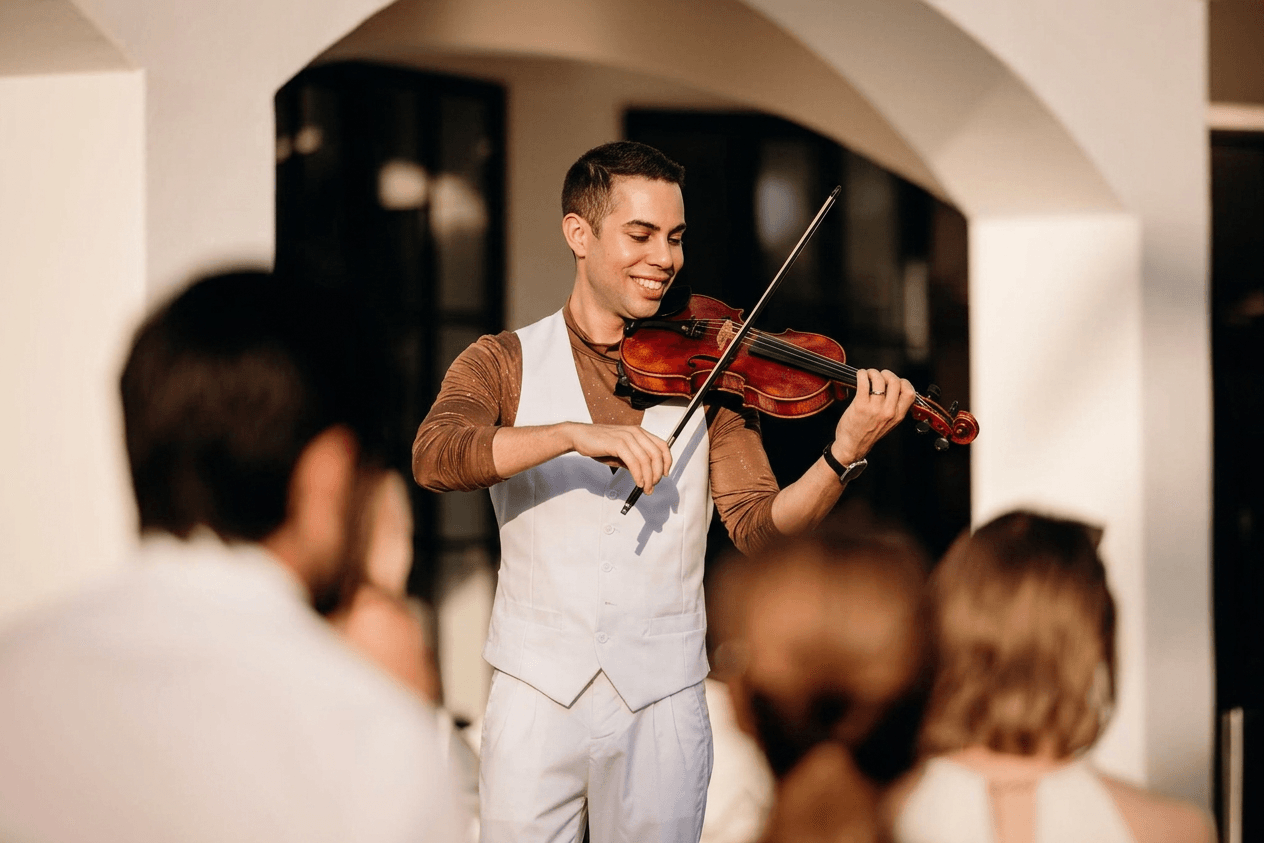 Young man playing violin in front of an audience indoors, smiling, with blurred people in foreground.