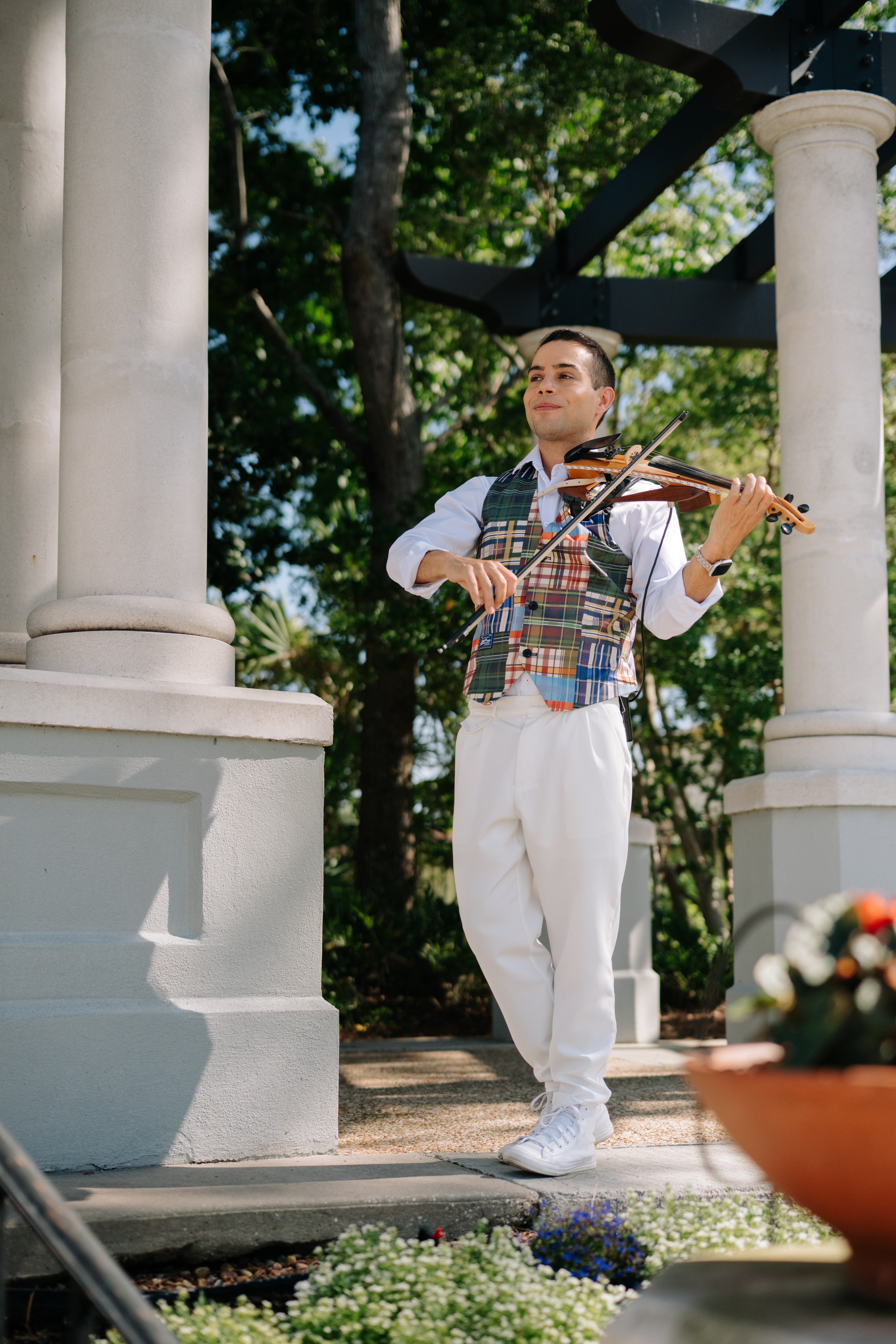 Young man playing violin outdoors between white columns, trees in background, sunlight, wearing white clothes and a plaid vest.