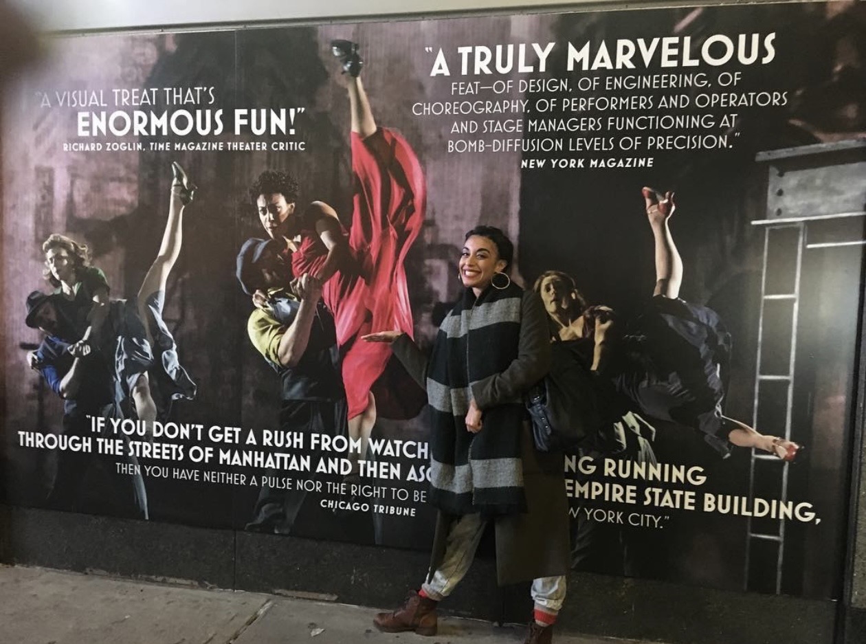 Three children stand in front of a large movie poster, smiling and raising their arms. The poster features people dancing and text about a Marvelous event.