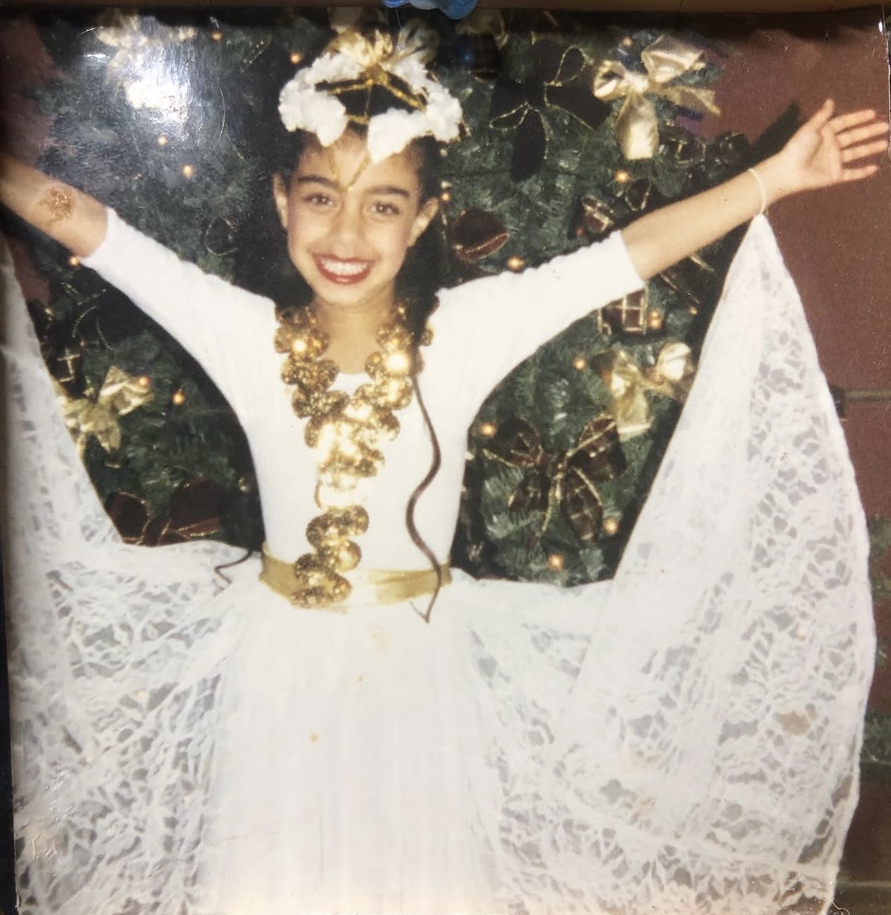 Young girl in a white dress with lace, smiling with arms outstretched, standing in front of a decorated Christmas tree.