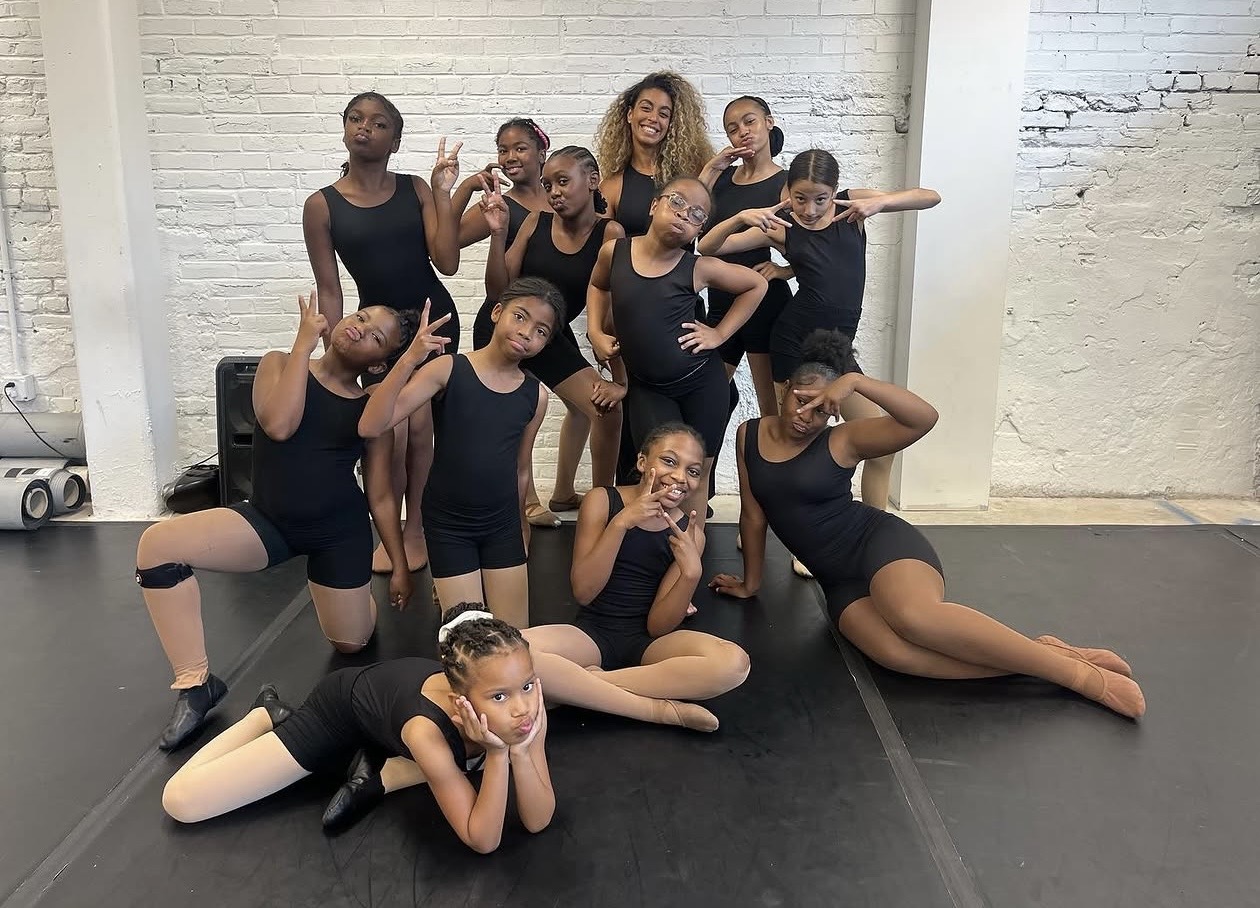 Group of young dancers in black costumes posing in a dance studio with white brick walls.