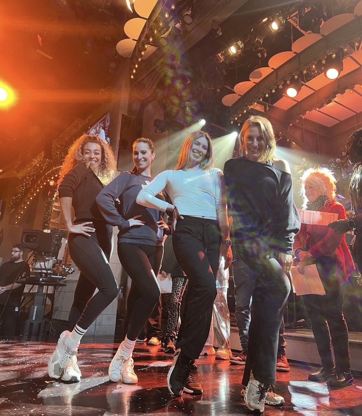 Four women dancing on stage with colorful lighting and a curved ceiling, smiling and wearing casual outfits with sneakers.