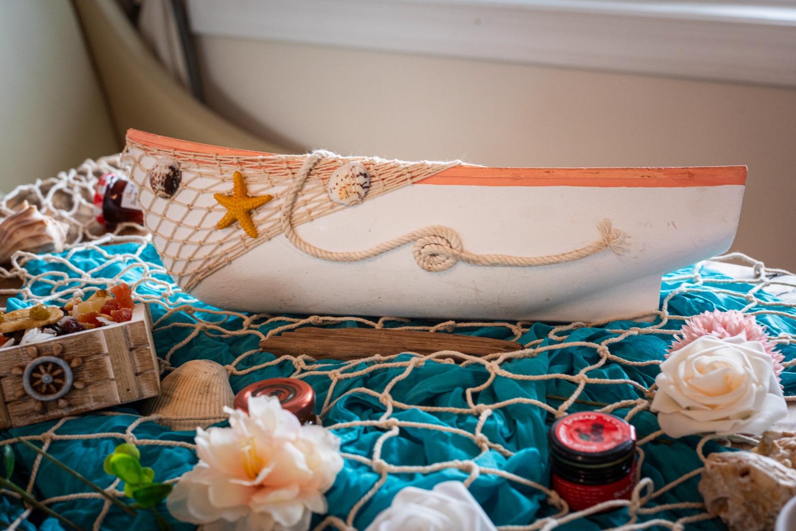 Decorative boat with starfish and rope, surrounded by flowers, jars, and a wooden box on a blue netted surface.