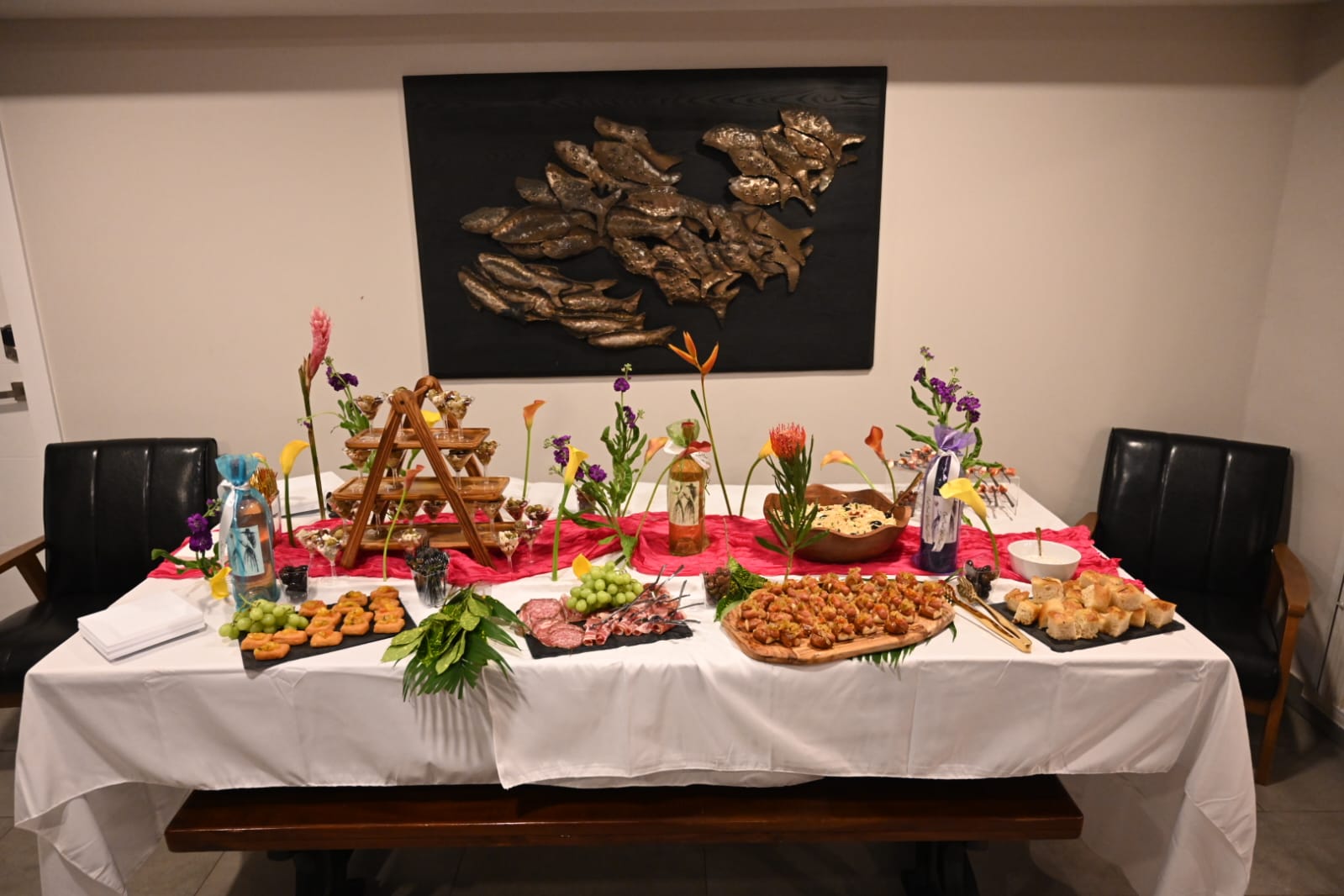 Table with food and decorations, black wall art above, chairs on sides, white tablecloth, colorful flowers, and various dishes.