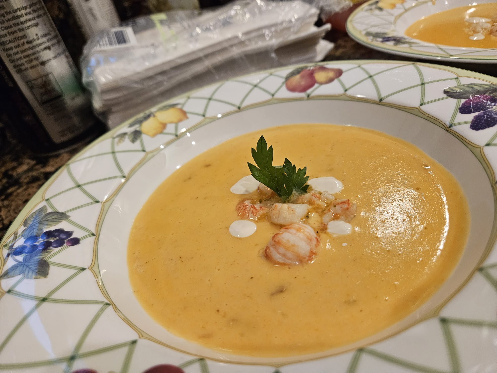 Bowl of creamy orange soup garnished with herbs and small toppings, on a decorative plate with floral pattern.