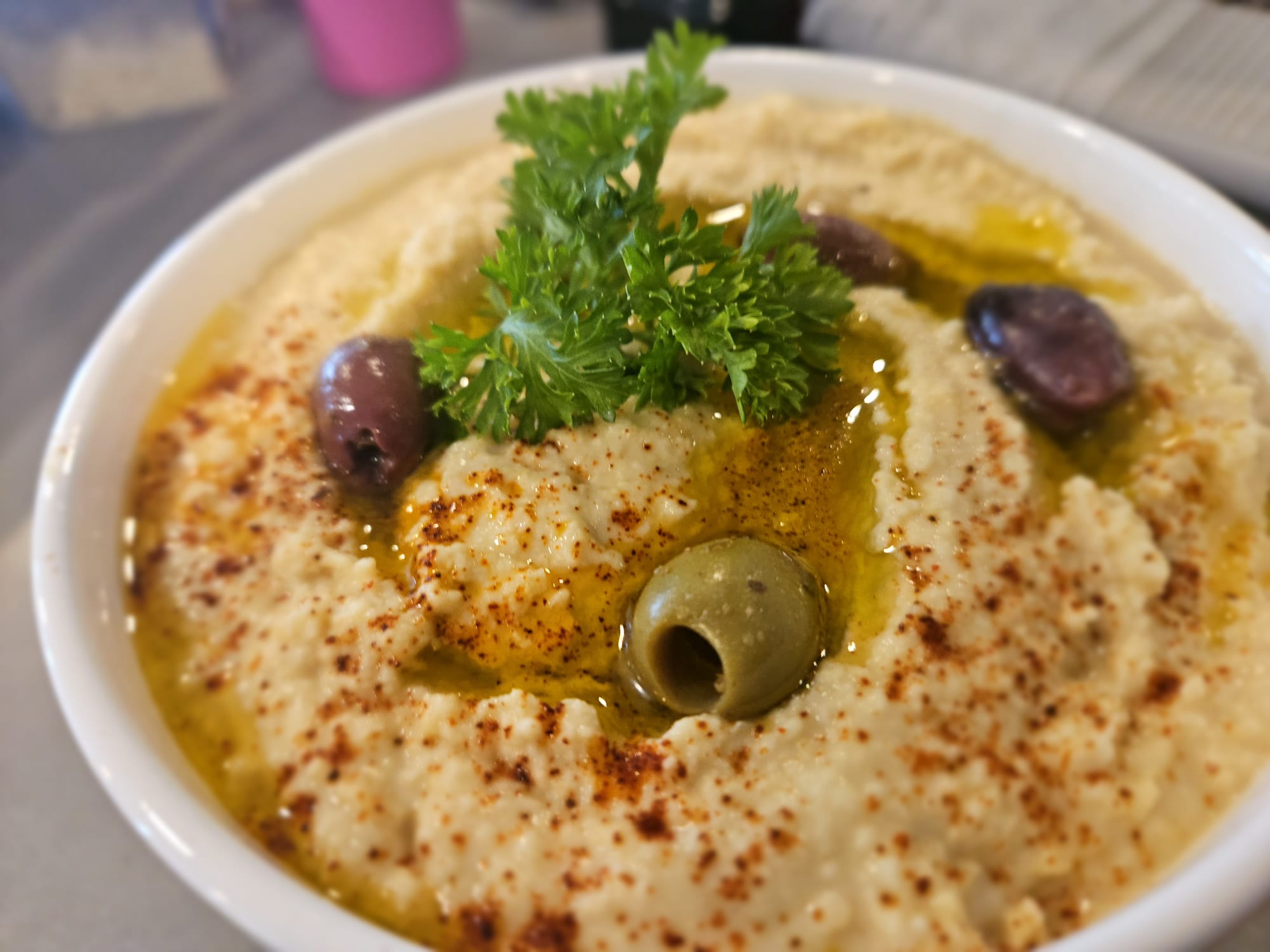 Bowl of hummus garnished with parsley, olives, and paprika, with a creamy texture and olive oil drizzle.