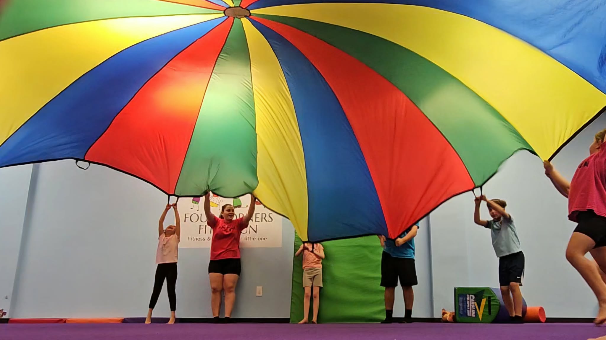 People holding a large multicolored parachute indoors, with a banner in the background, and a purple floor.