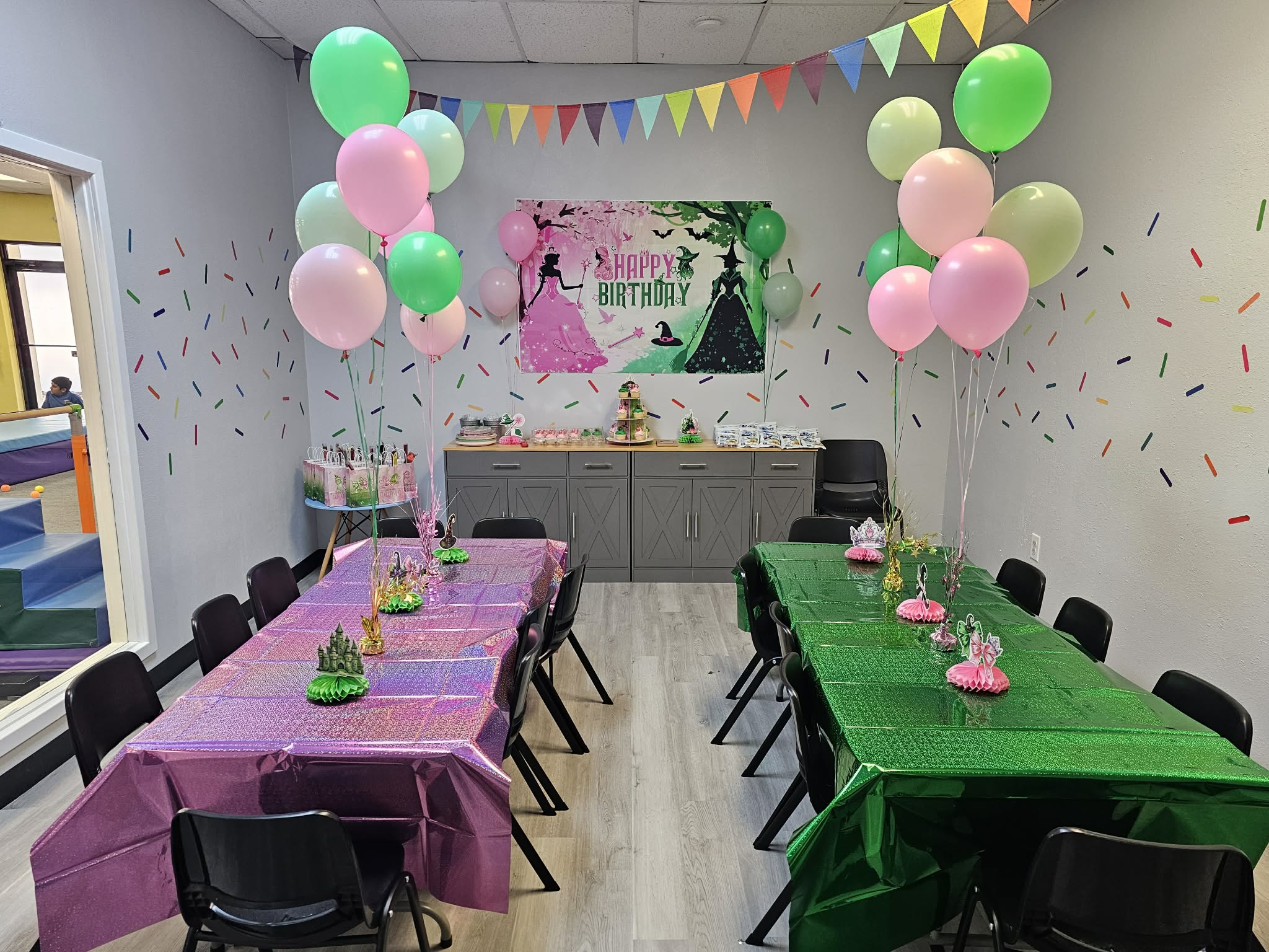 Party room decorated with balloons, colorful banners, and tables with pink and green tablecloths for a birthday celebration.