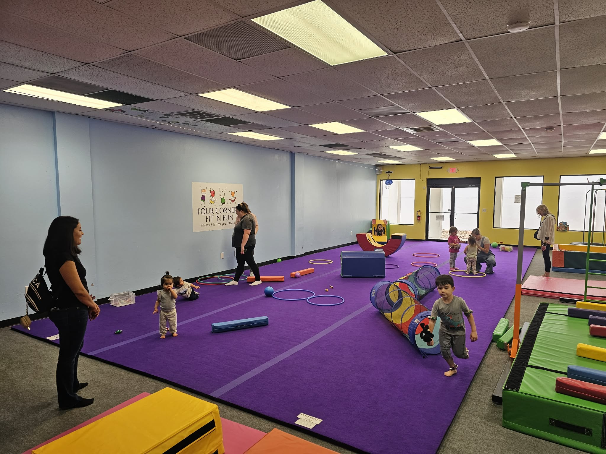 Children and adults in a playroom with purple carpet, play equipment, and large windows, engaging in activities.