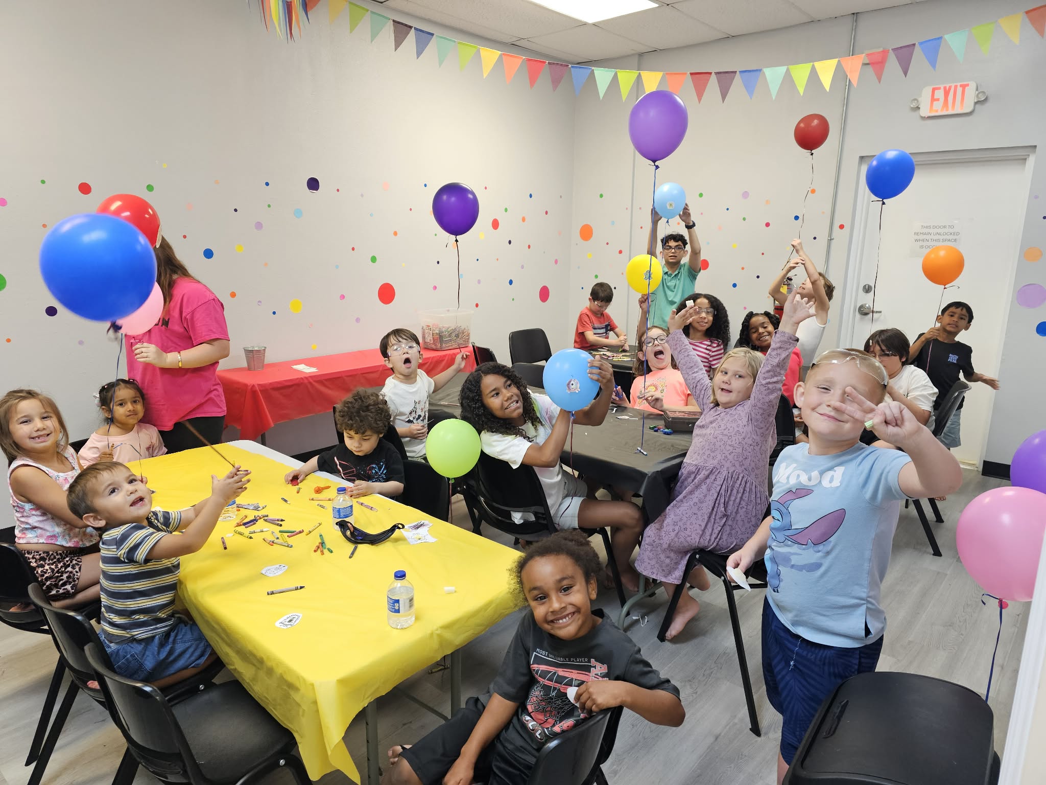 Children at a party with colorful balloons, decorations, and a table with a yellow tablecloth, smiling and playing.