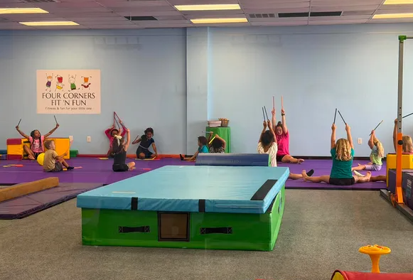 Children participate in a gymnastics class on mats, with a blue foam pit in the foreground and a sign on the wall.