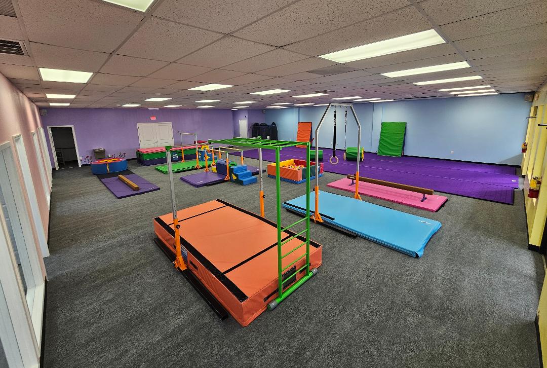Indoor gym with mats, bars, and climbing equipment in a spacious room with purple and blue walls.