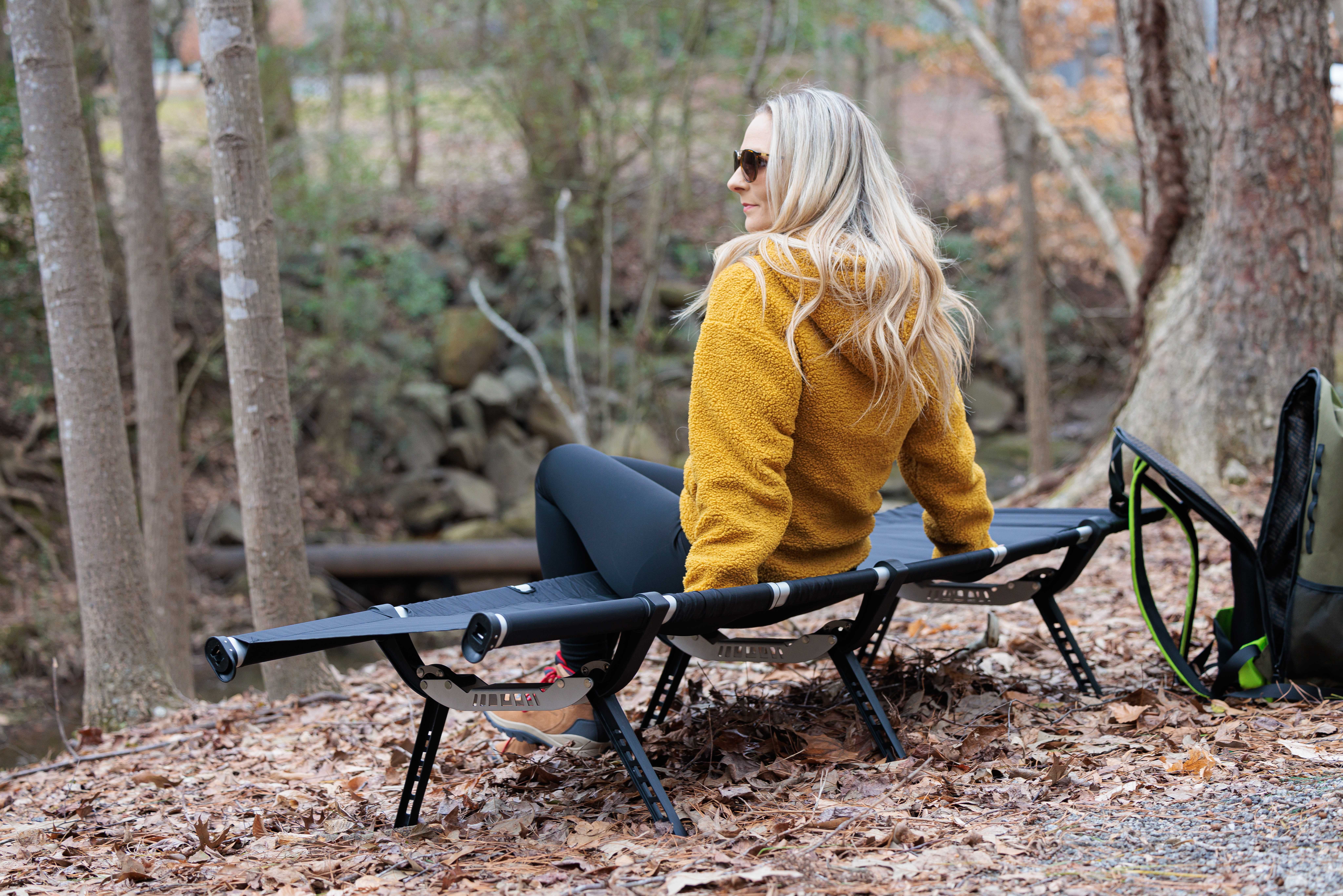 Woman with long blonde hair wearing a yellow jacket sitting on a black bench outdoors, surrounded by trees and fallen leaves.