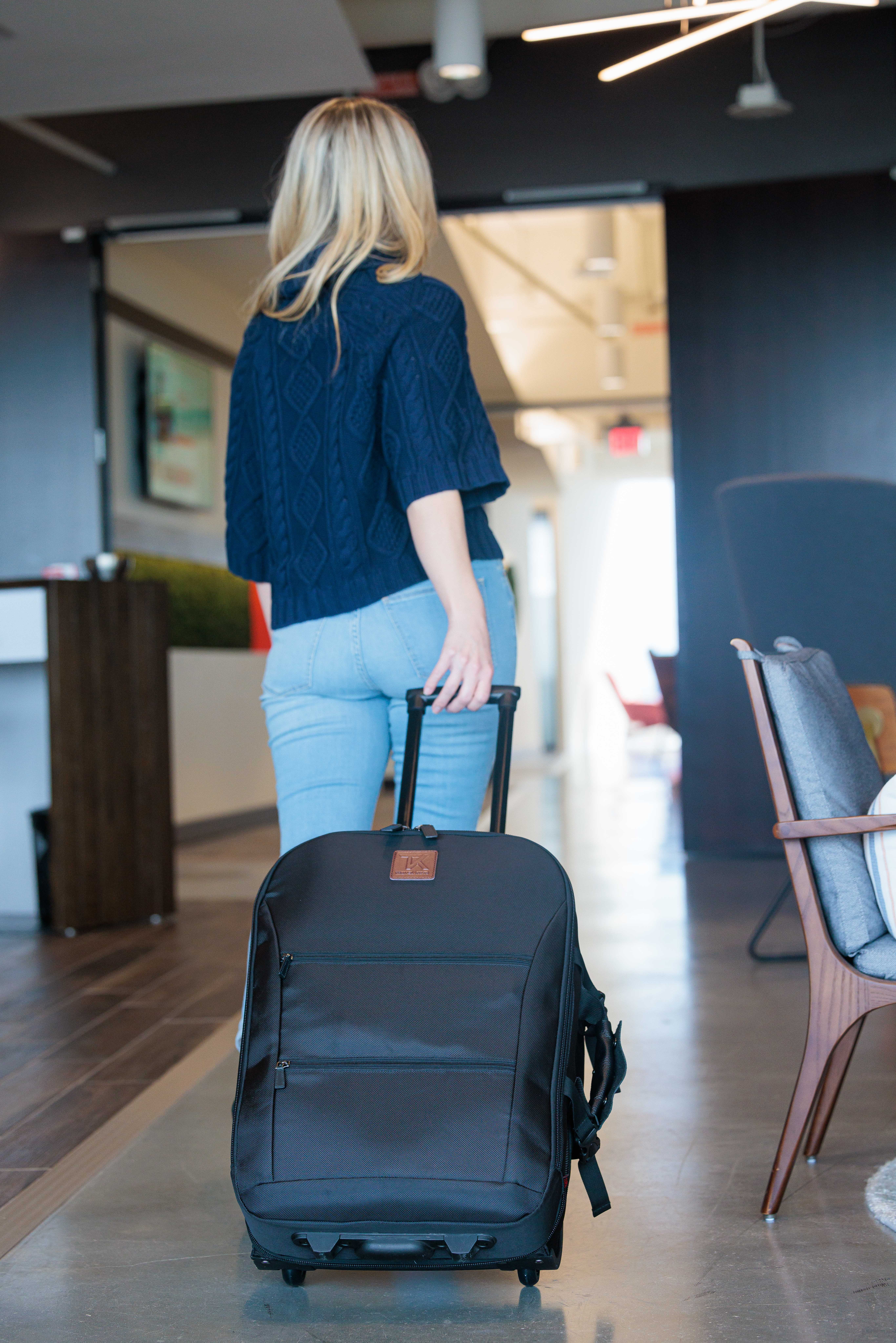 Woman with blonde hair, wearing a blue sweater and light blue pants, pulls a black rolling suitcase in a modern indoor space.