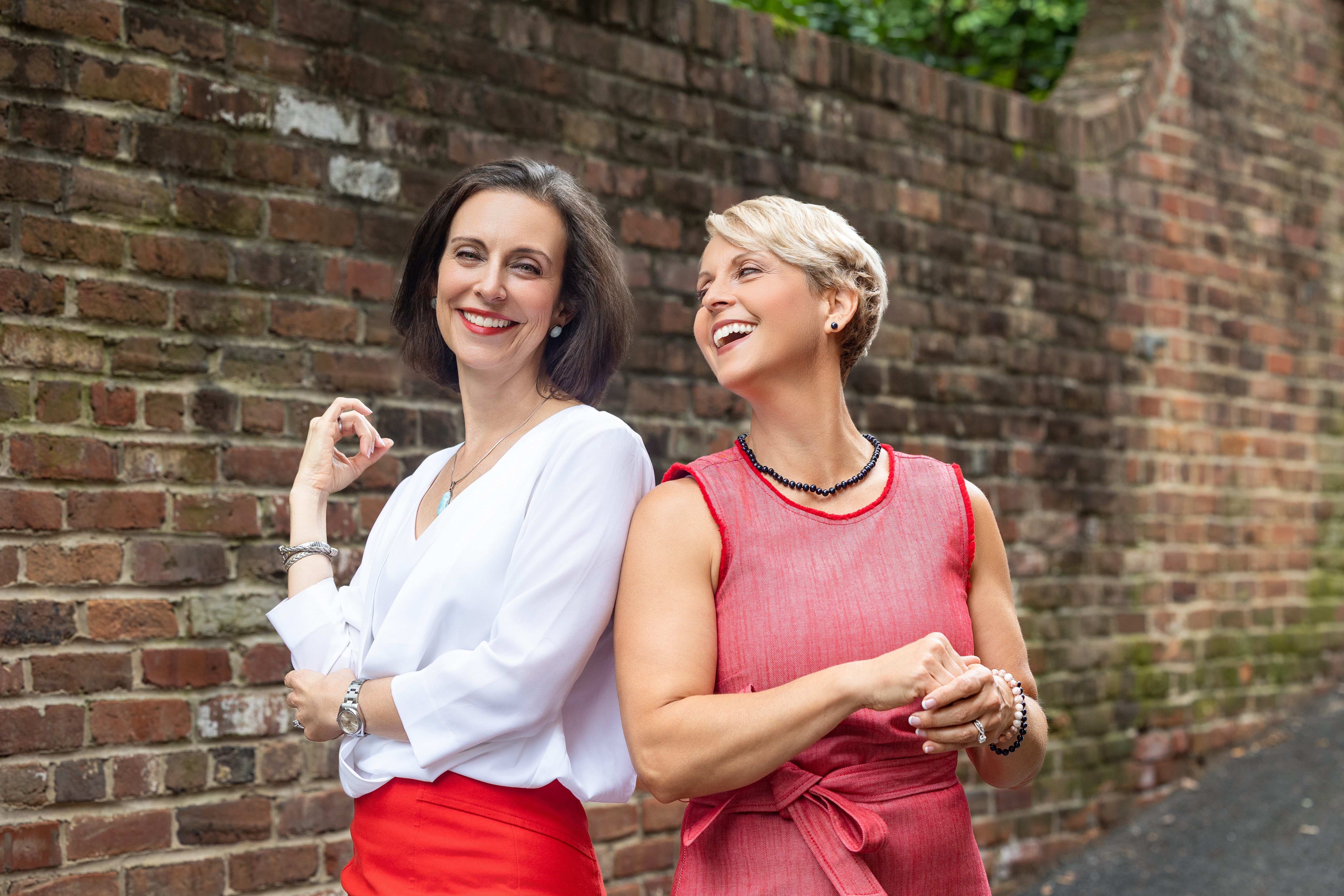 Two women smiling and talking outdoors near a brick wall, one wearing a white top and red skirt, the other in a red sleeveless dress.