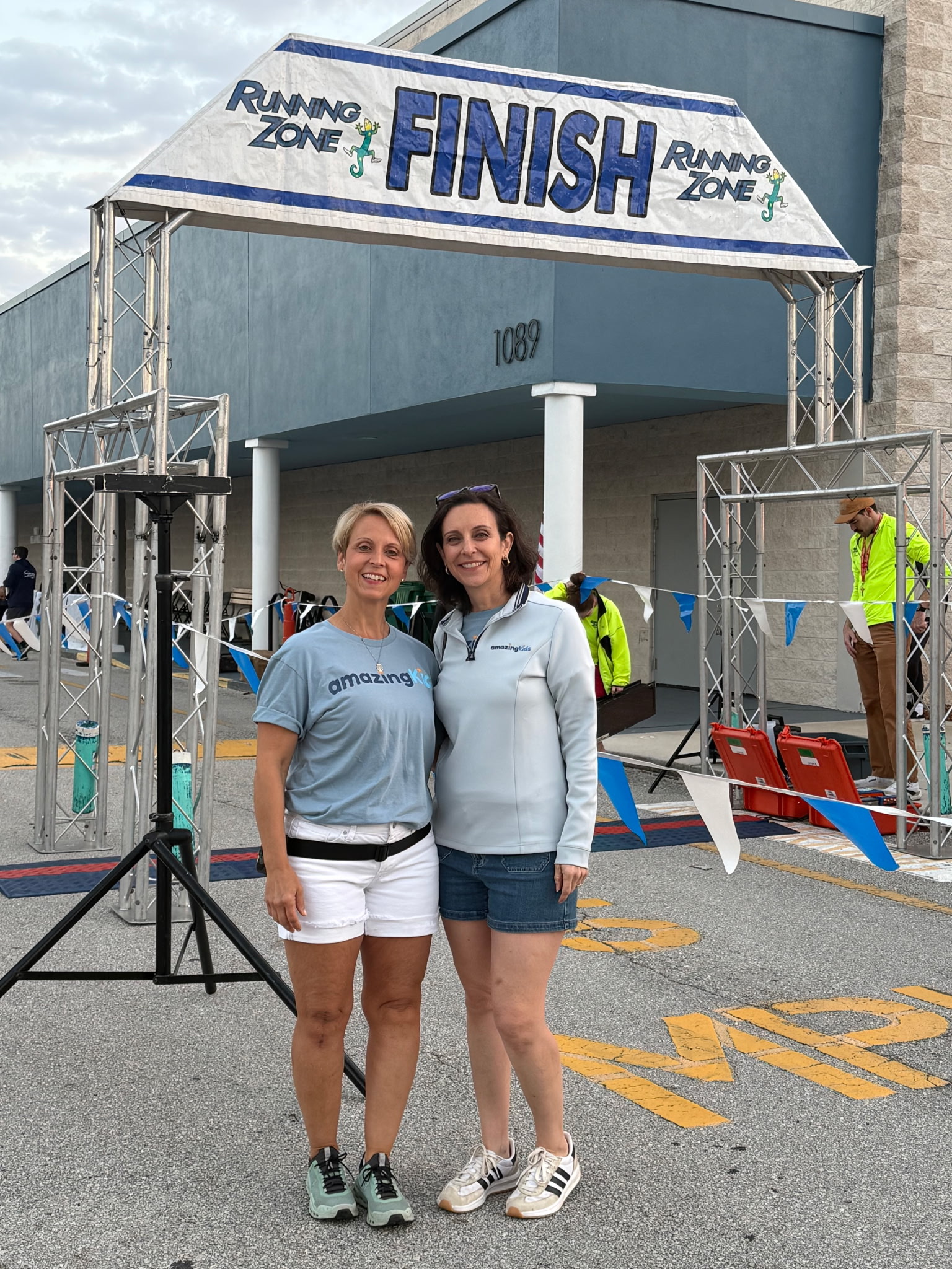 Two women standing in front of a finish line banner at a race event, smiling, with people and a building in the background.