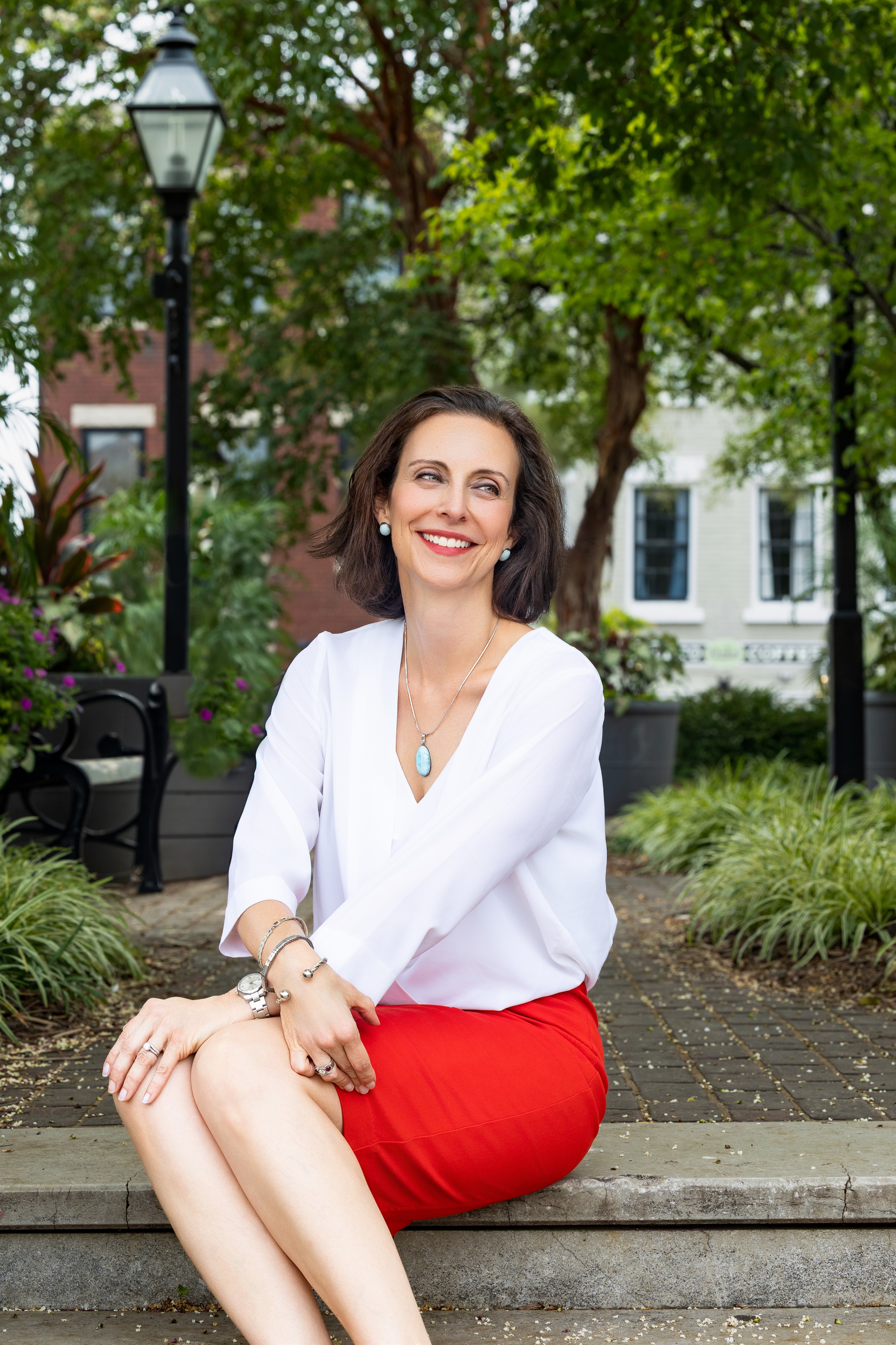 Woman sitting on a park bench smiling, wearing a white blouse and red skirt, with trees and buildings in background.