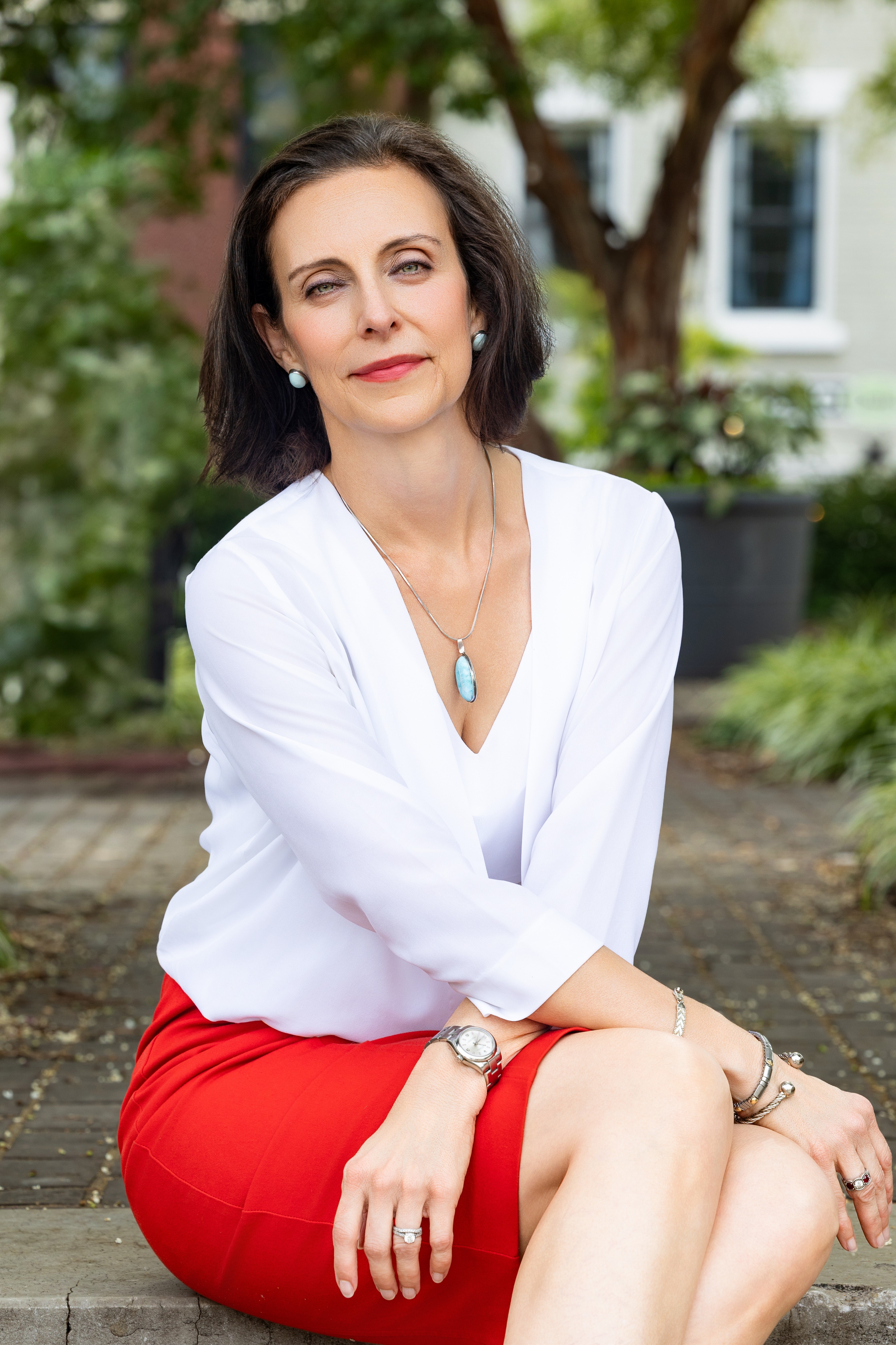Woman with dark hair in a white blouse and red skirt sitting outdoors, with trees and buildings in background.