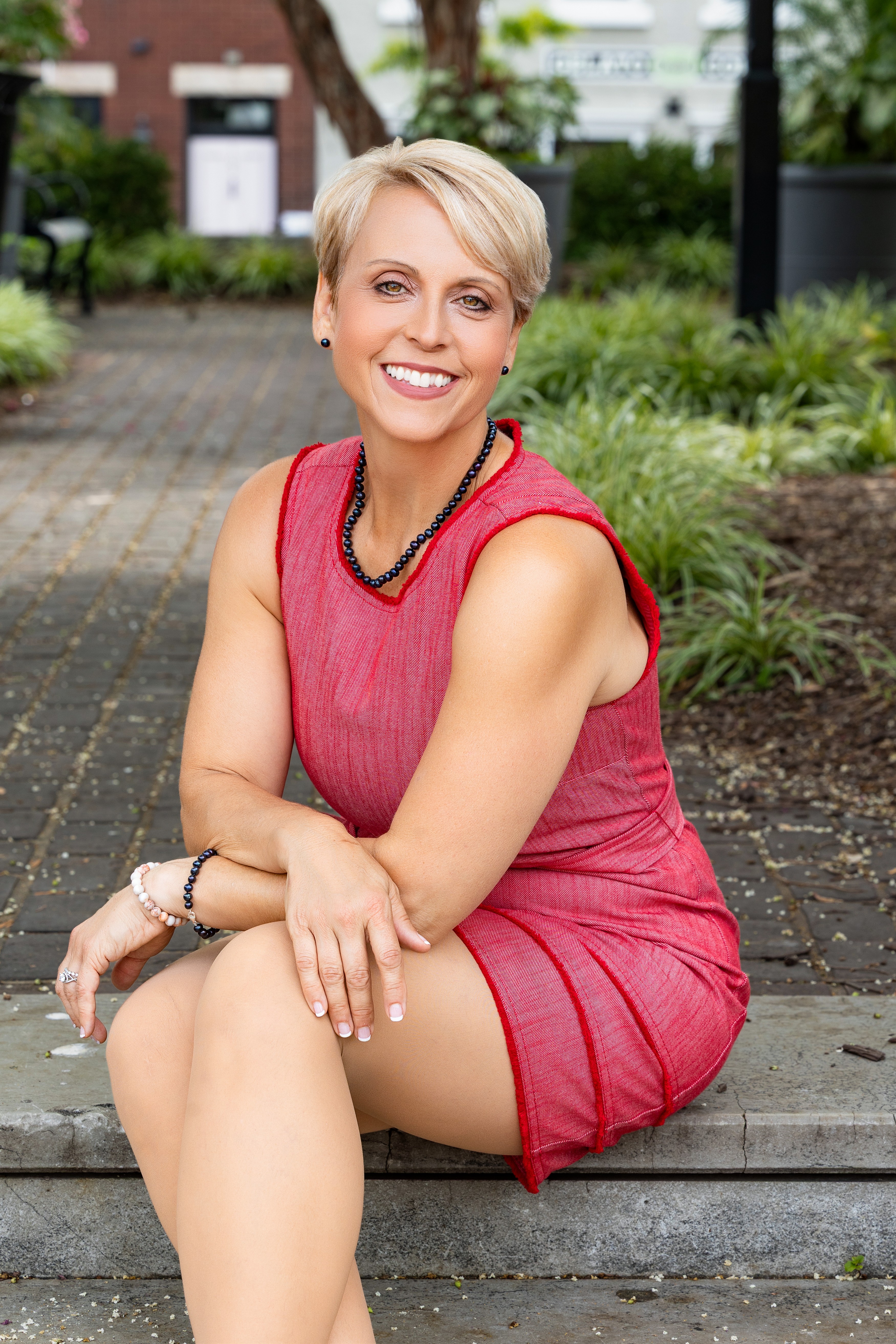 Smiling woman with short blonde hair in a red sleeveless dress sitting on a park bench, surrounded by greenery.