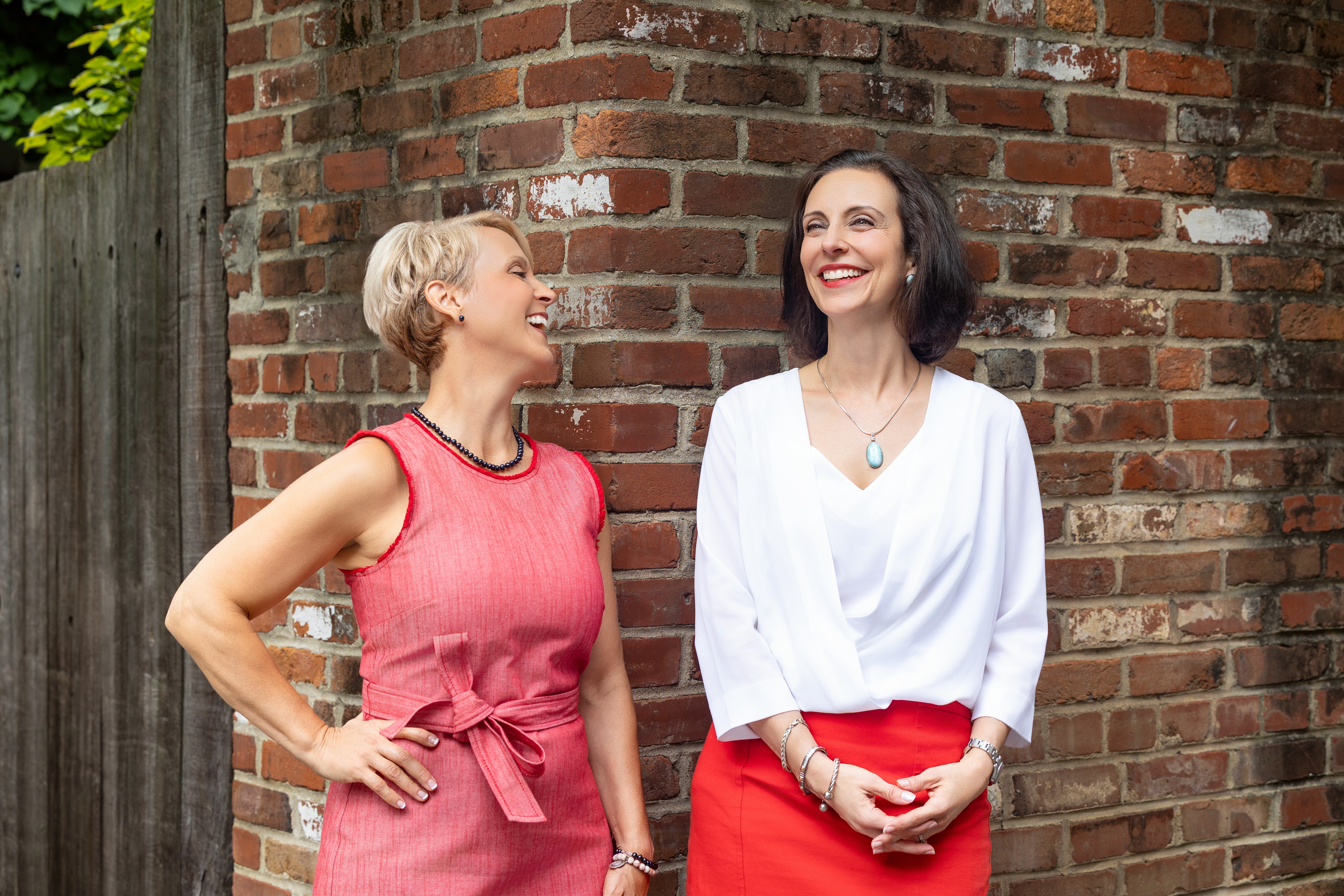 Two women standing outdoors in front of a brick wall, smiling and talking, one wearing a pink dress, the other in a white top and red skirt.