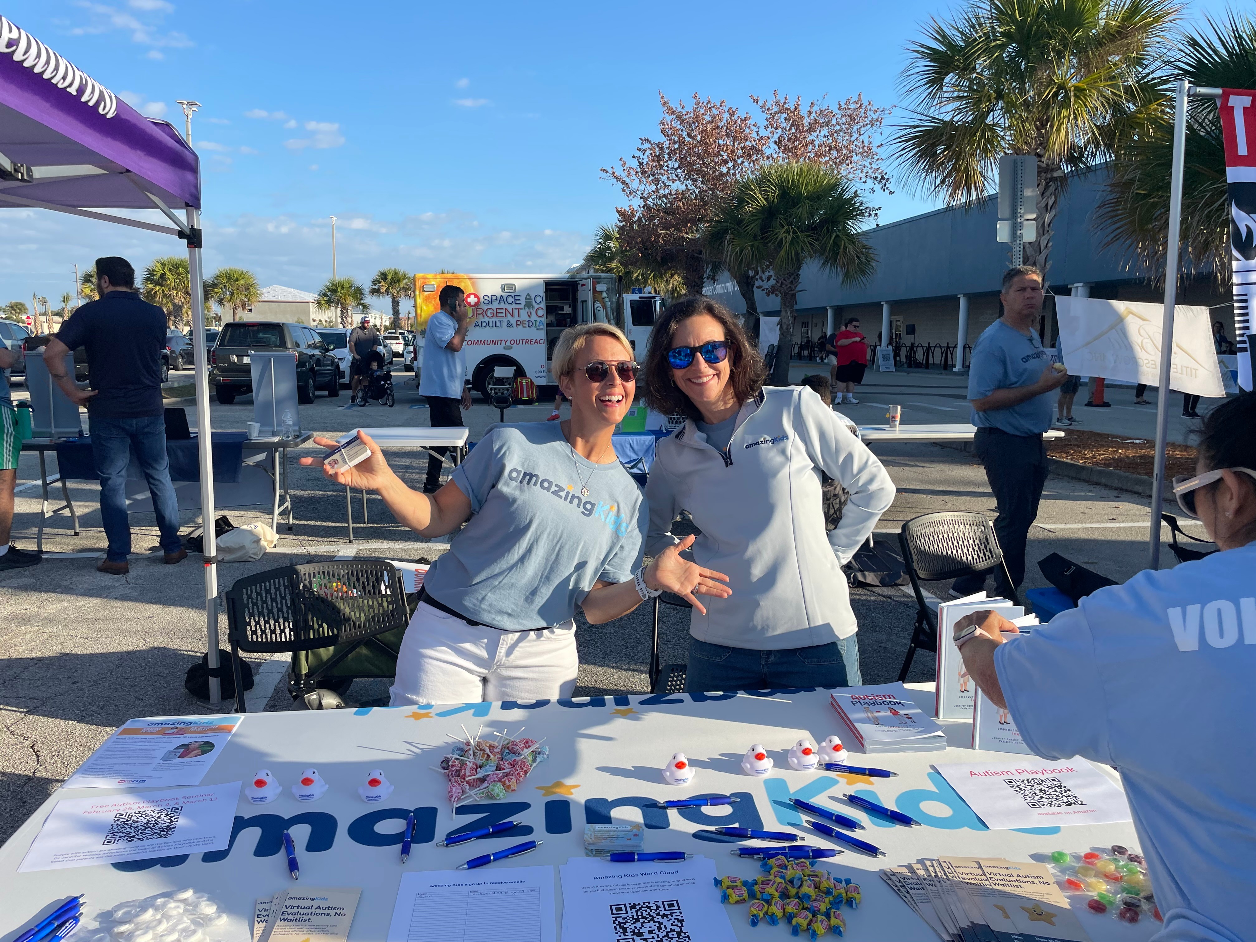 Two women smiling and posing at an outdoor event table with a purple canopy, palm trees, and people in the background.