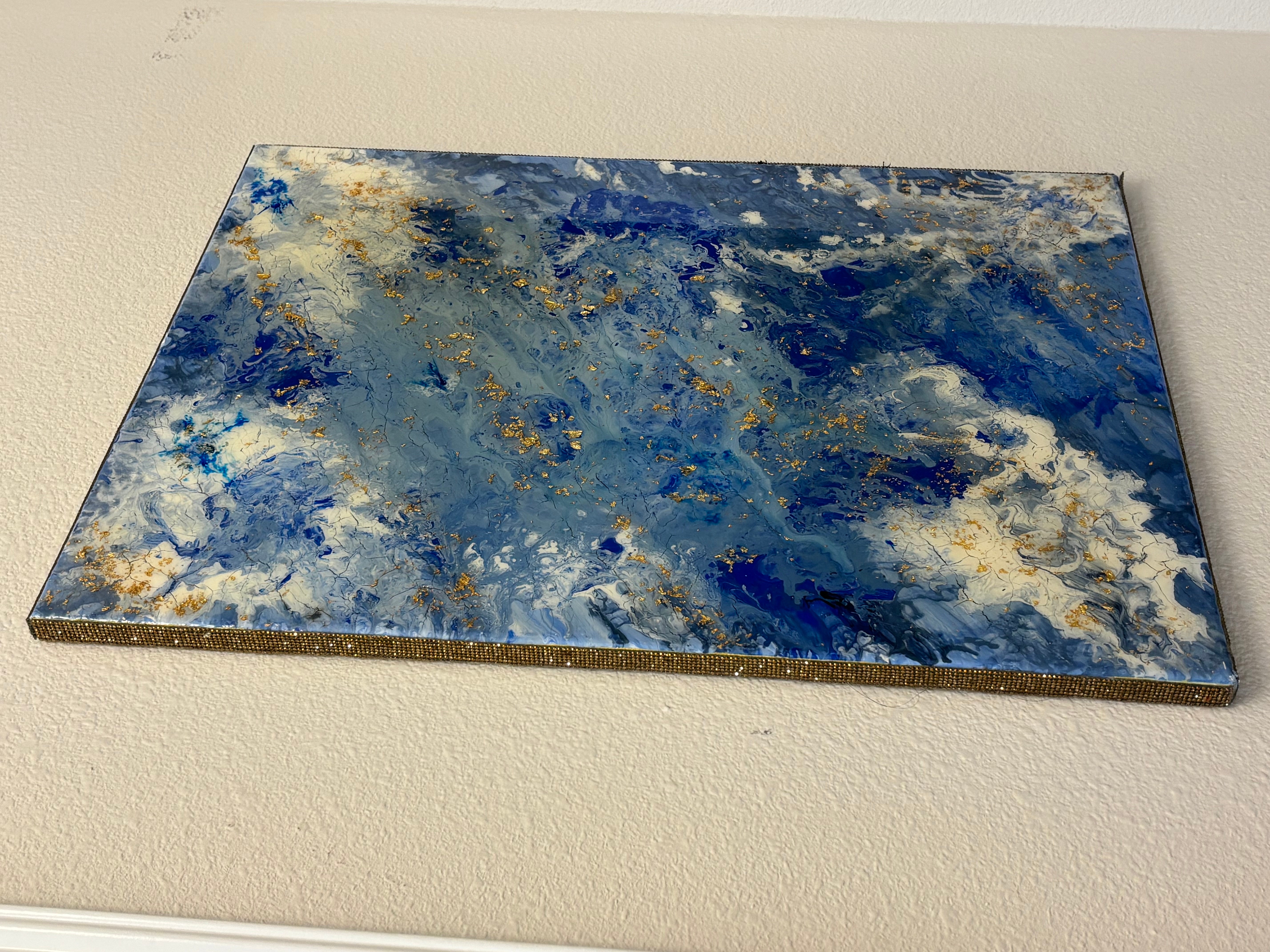 Abstract painting with blue, white, and gold colors on a rectangular canvas, hung on a wall.