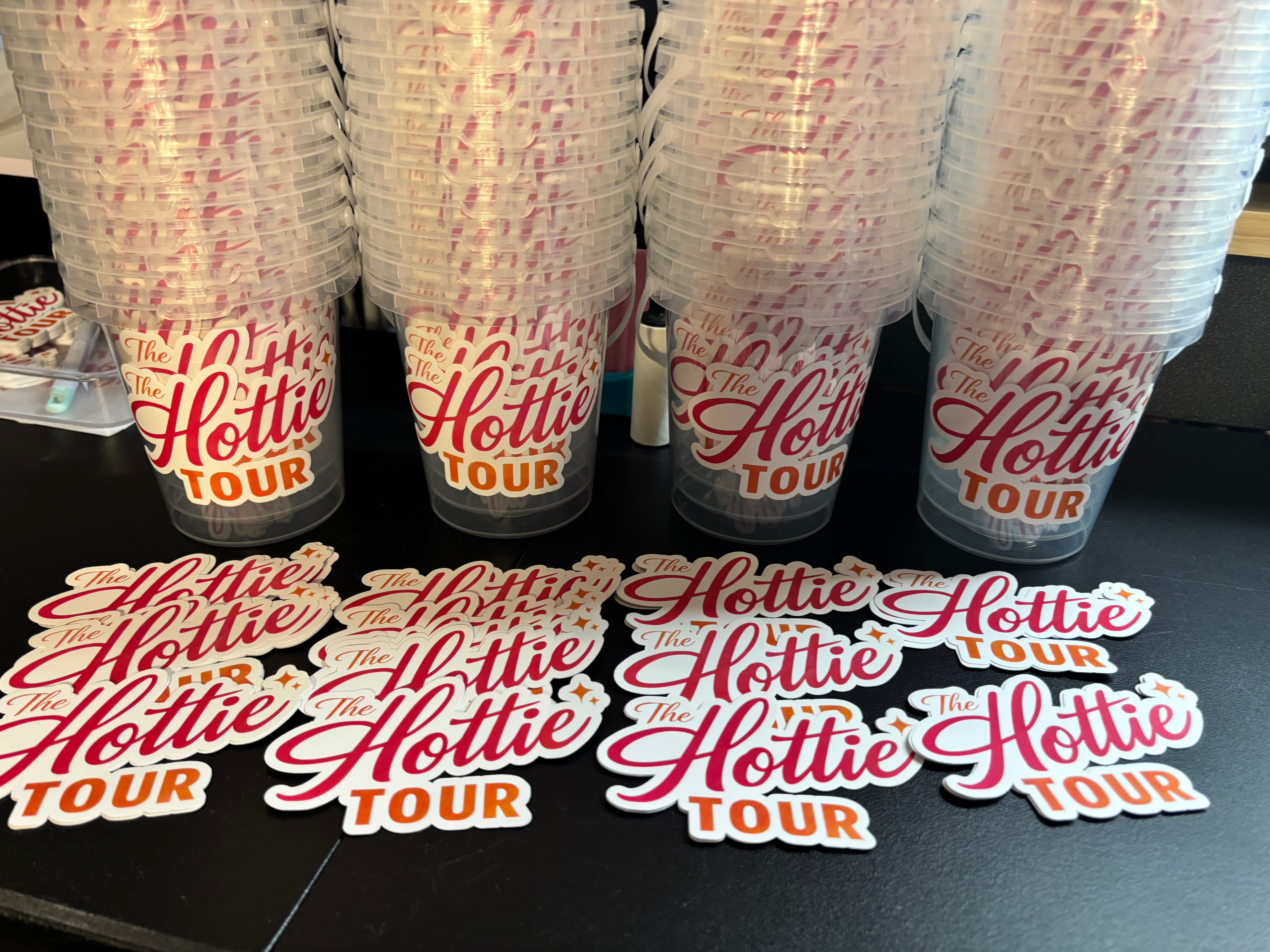 Six cups with red and black text on a black surface, with stickers reading 'Hottie Tour' in front.
