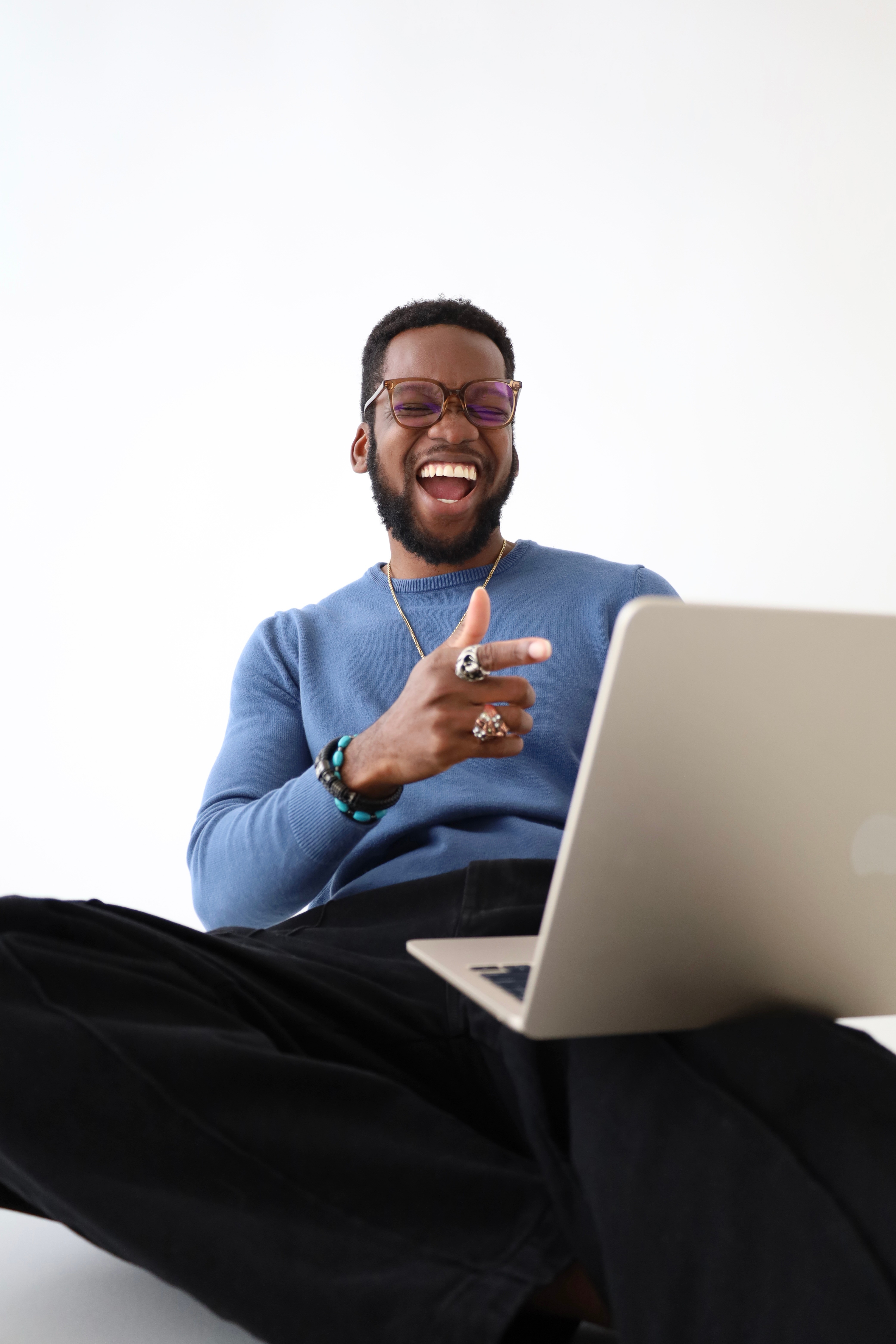 Person smiling and pointing at a laptop, wearing glasses and a blue sweater, sitting against a white background.