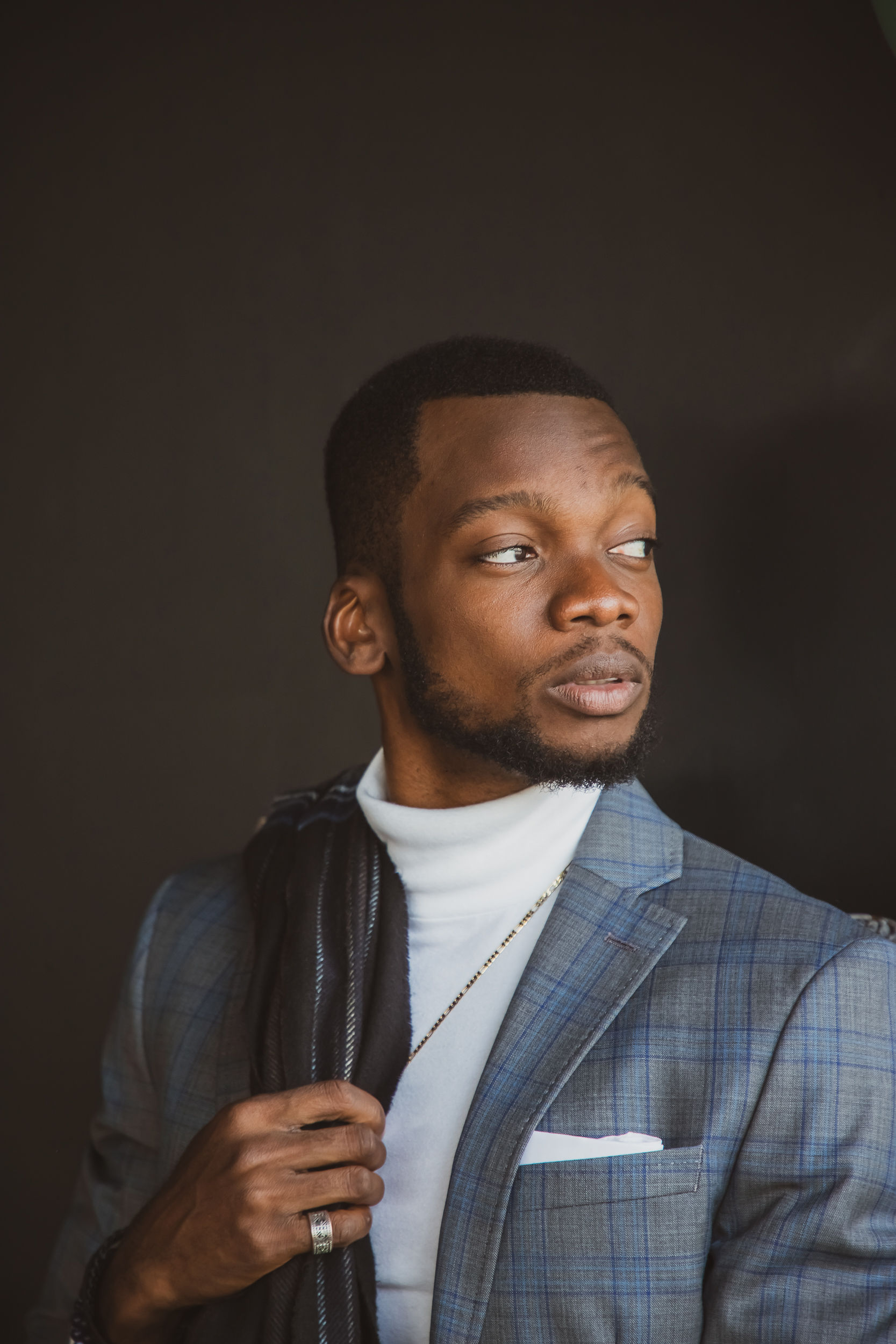 Young man in a plaid suit with a white turtleneck, looking to the side, against a dark background.