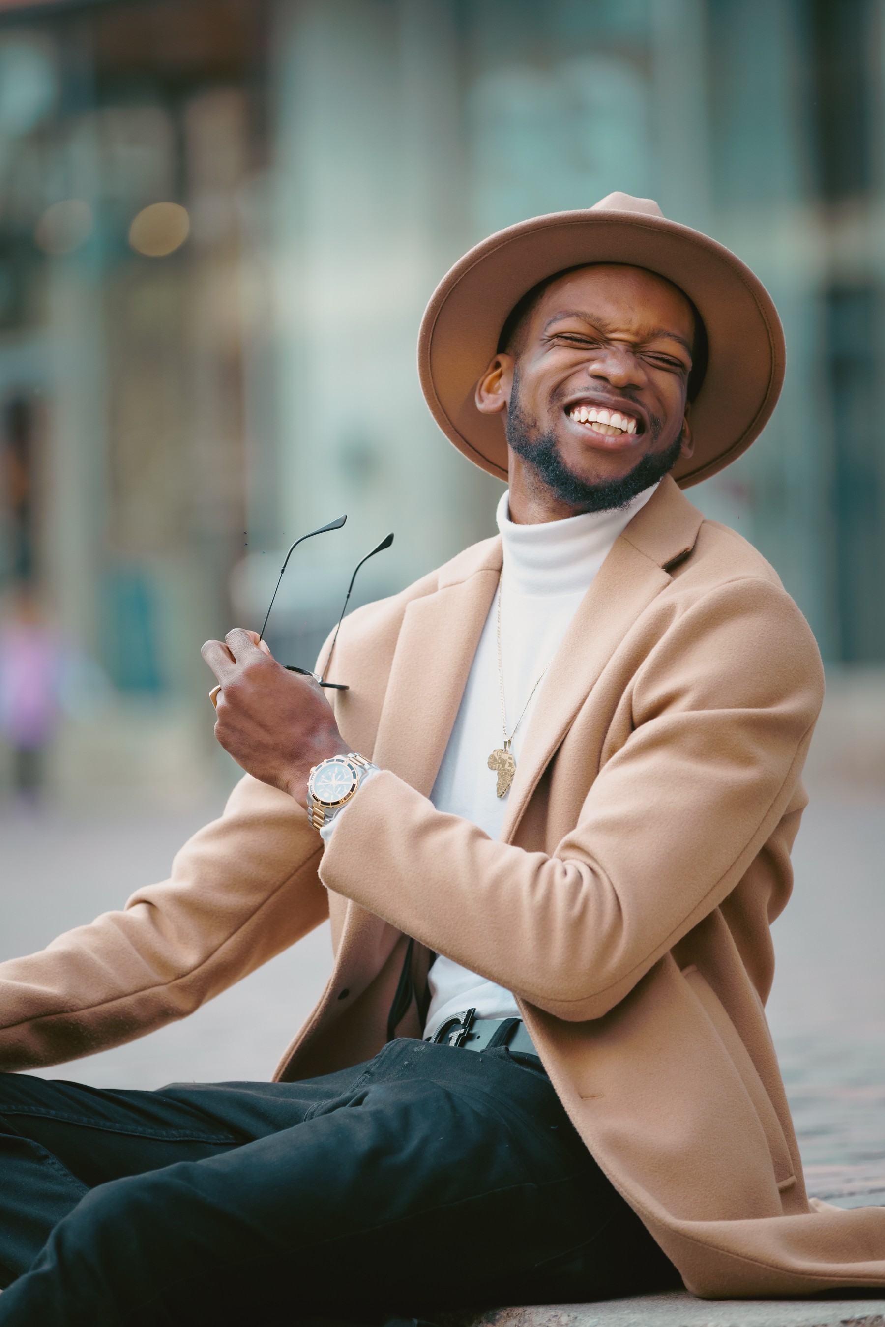 Smiling man wearing a hat and beige blazer, sitting outdoors with a blurred city background