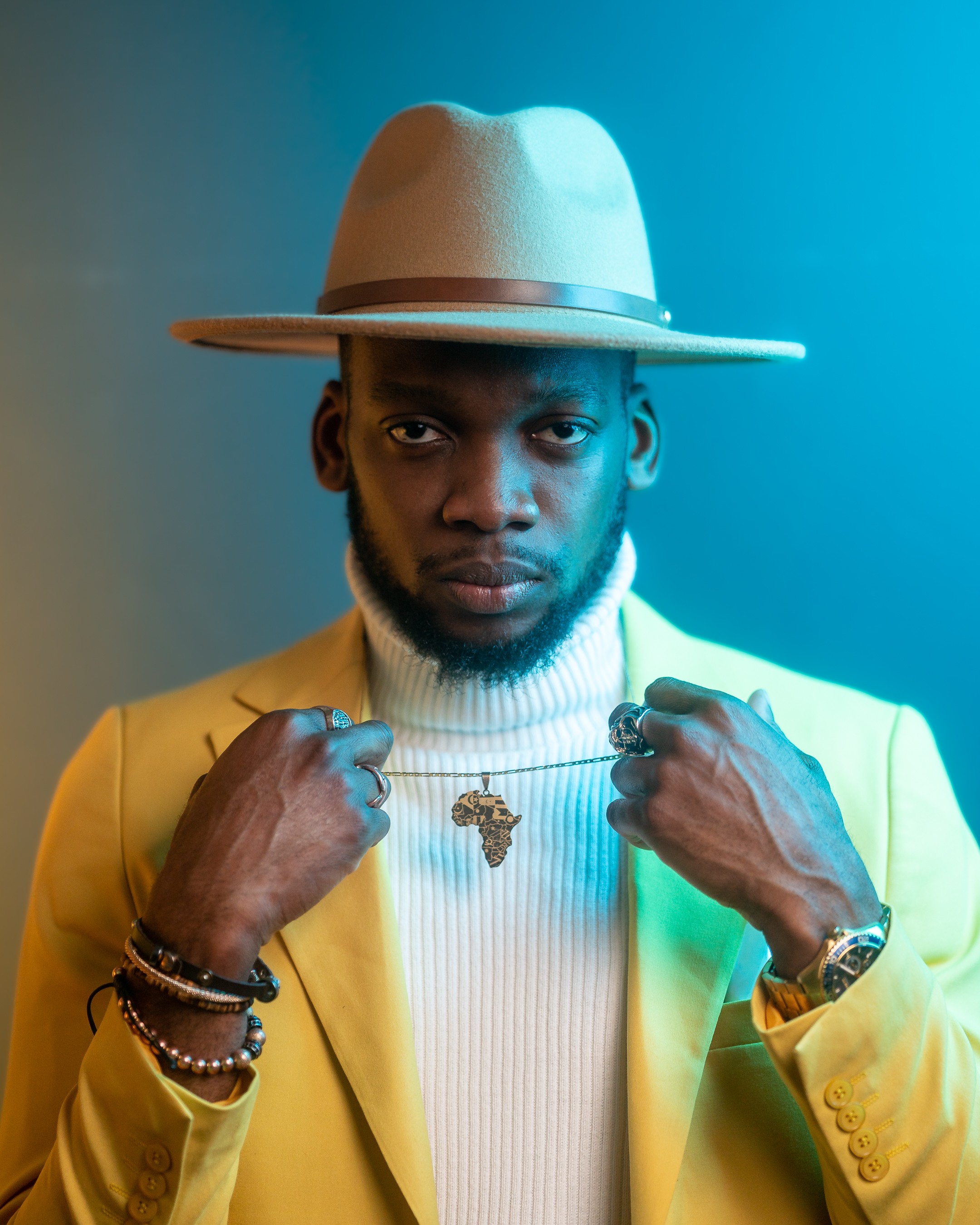 Man wearing a beige hat and yellow suit, holding a necklace with an African continent pendant, looking at the camera.