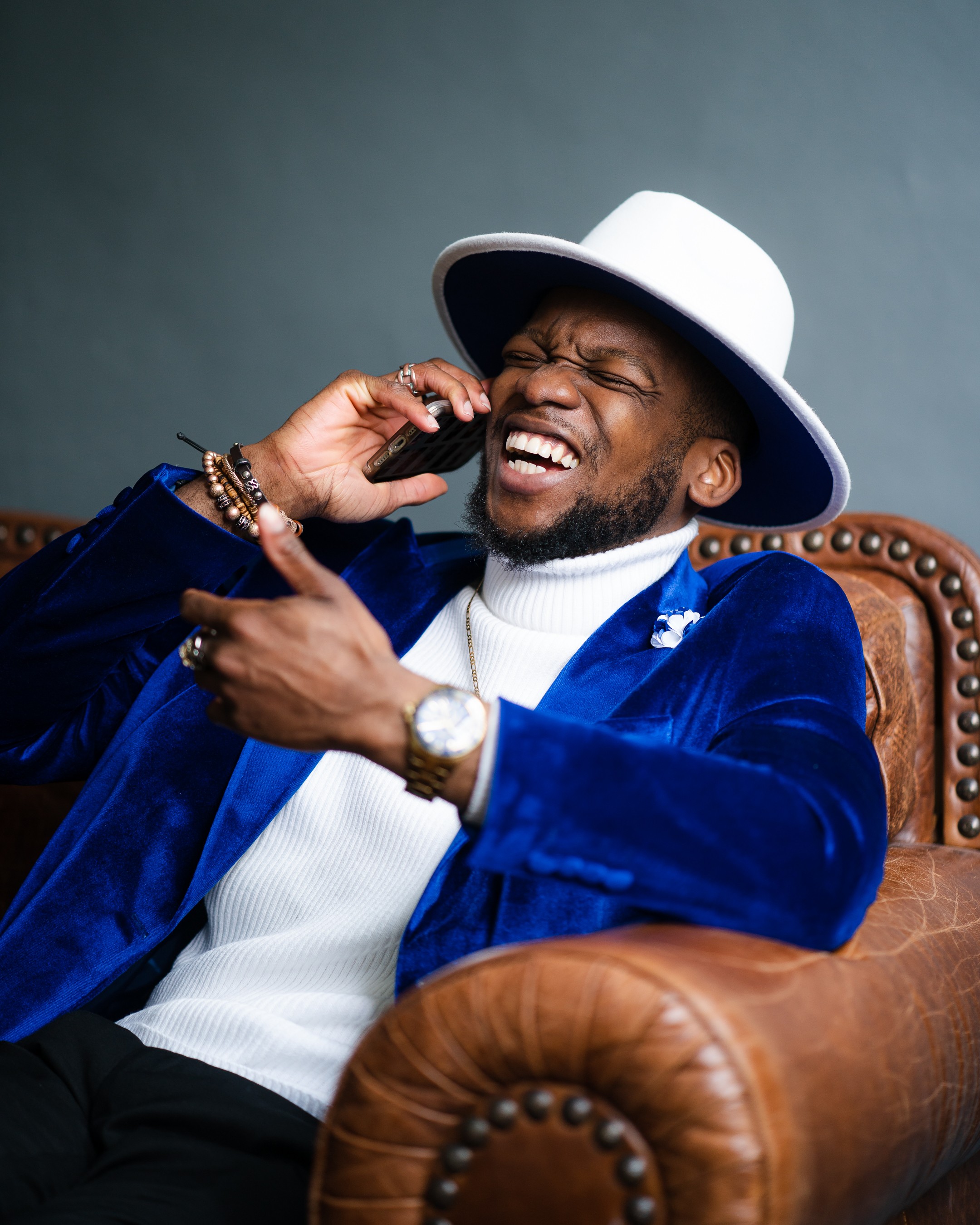 Man with a white hat and blue velvet jacket laughs while talking on a phone, sitting on a leather chair.