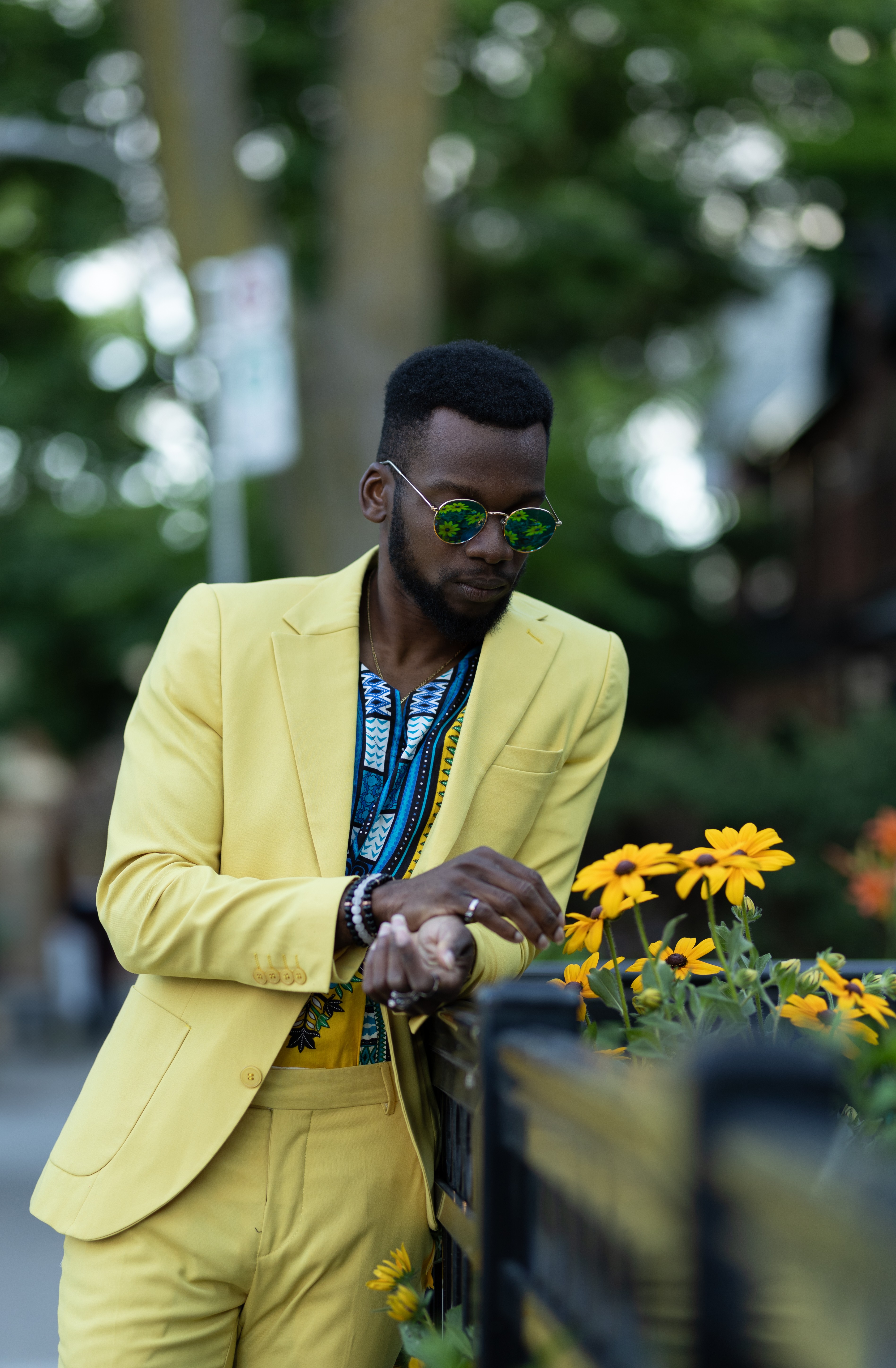 Person in yellow suit with sunglasses looking at yellow flowers outdoors, trees in background.