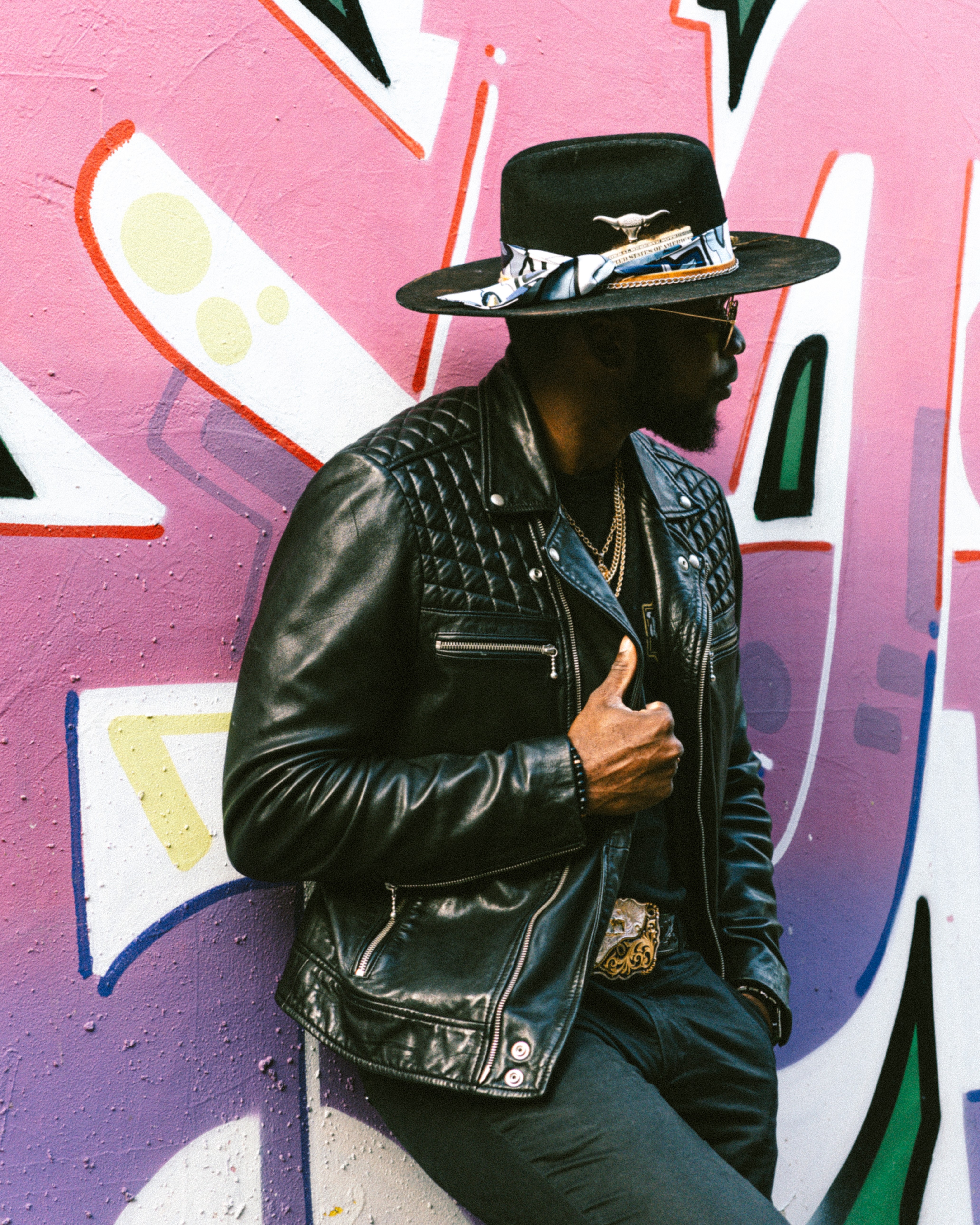 Person wearing a wide-brimmed hat, leather jacket, and sunglasses, standing against a colorful graffiti wall.
